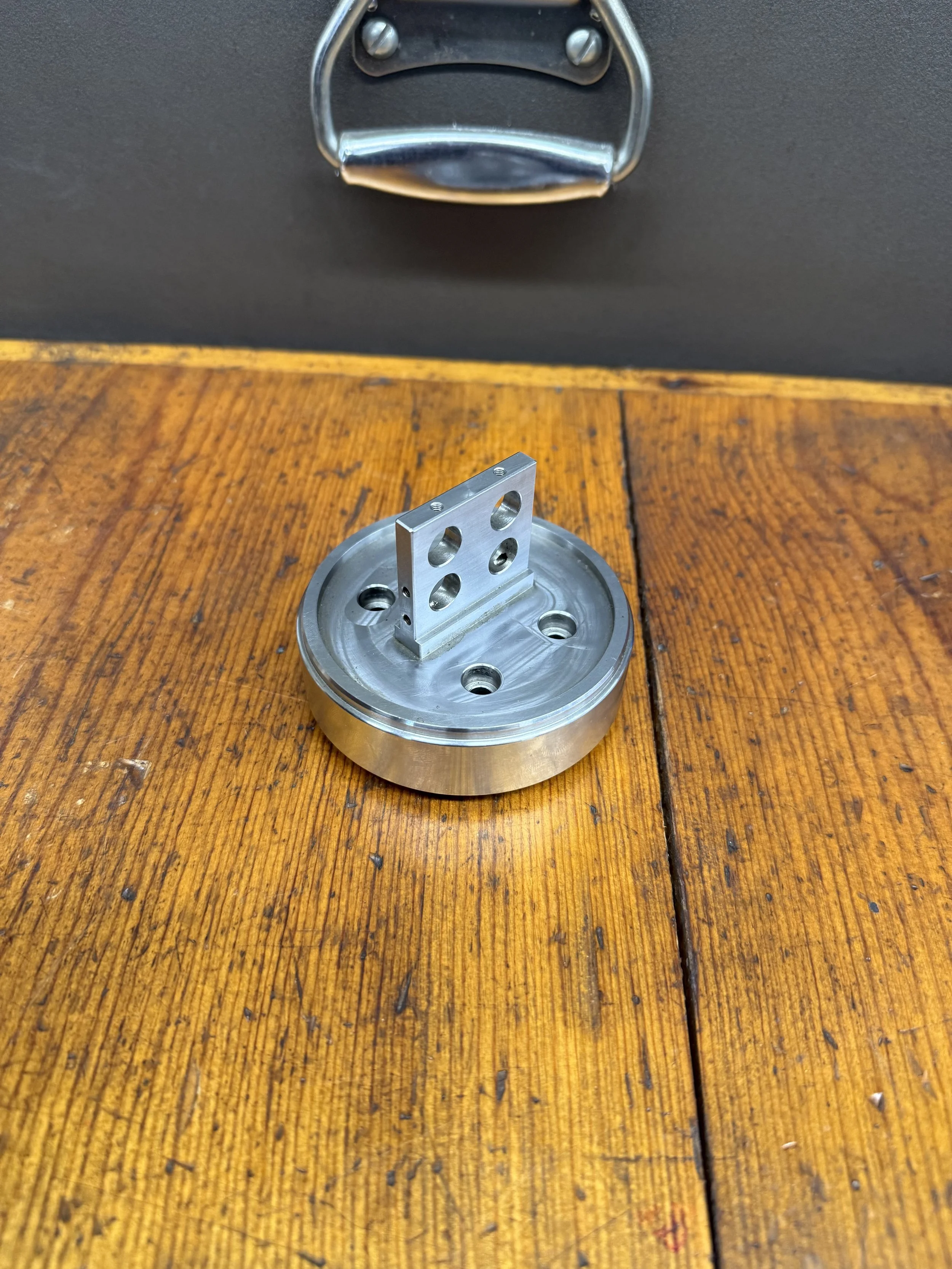 Metal mechanical component with a circular base and a vertical bracket with holes, placed on a wooden surface.