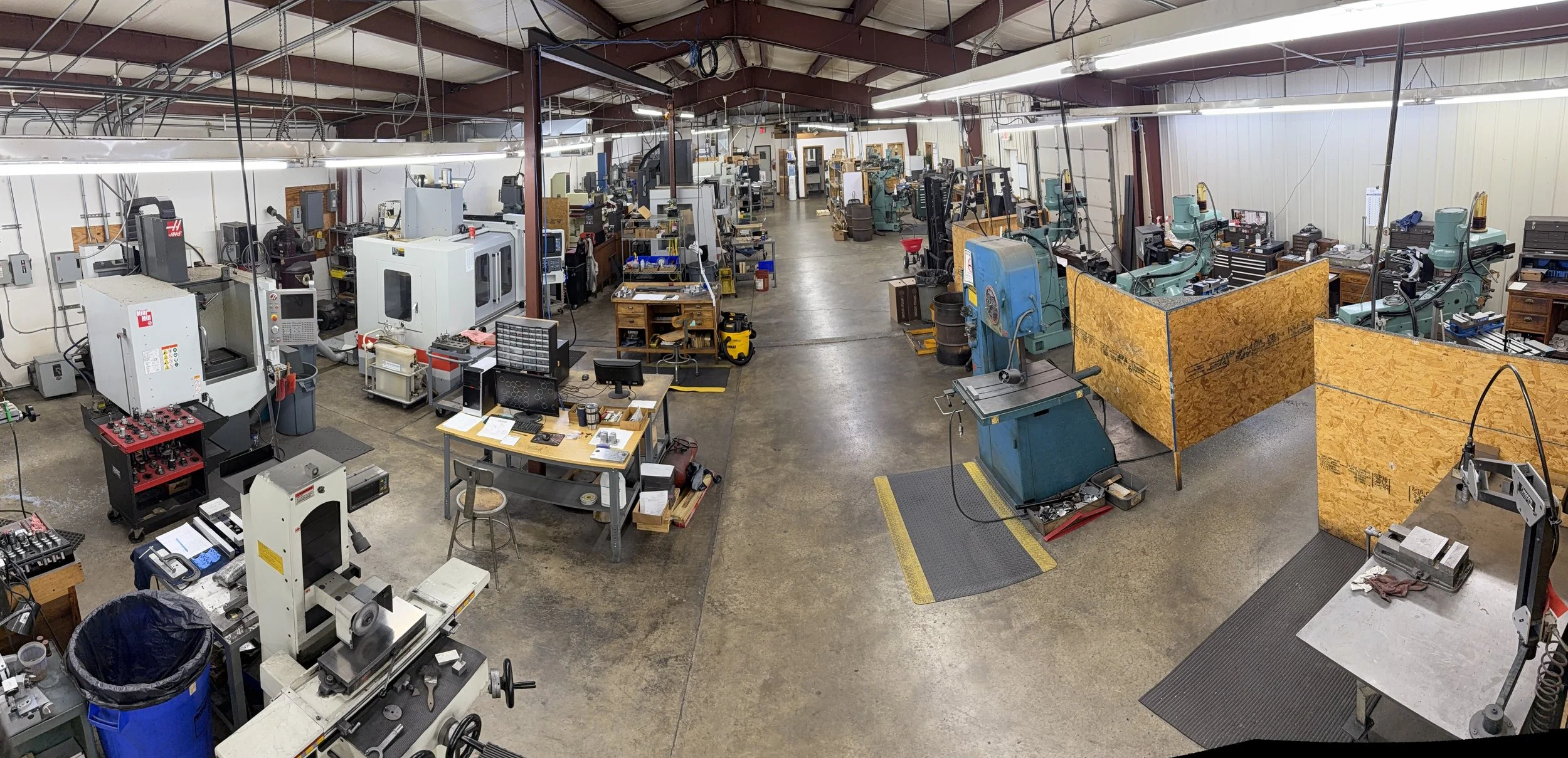 A spacious industrial workshop with various CNC machines, workbenches, and tools. The area is lit with overhead lighting, and there are partitions and safety mats near machines.
