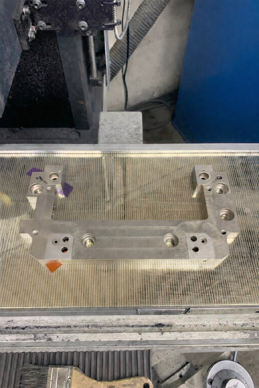 Metal machining fixture or mold part with multiple holes and a rectangular cutout, placed on a workbench in a workshop.