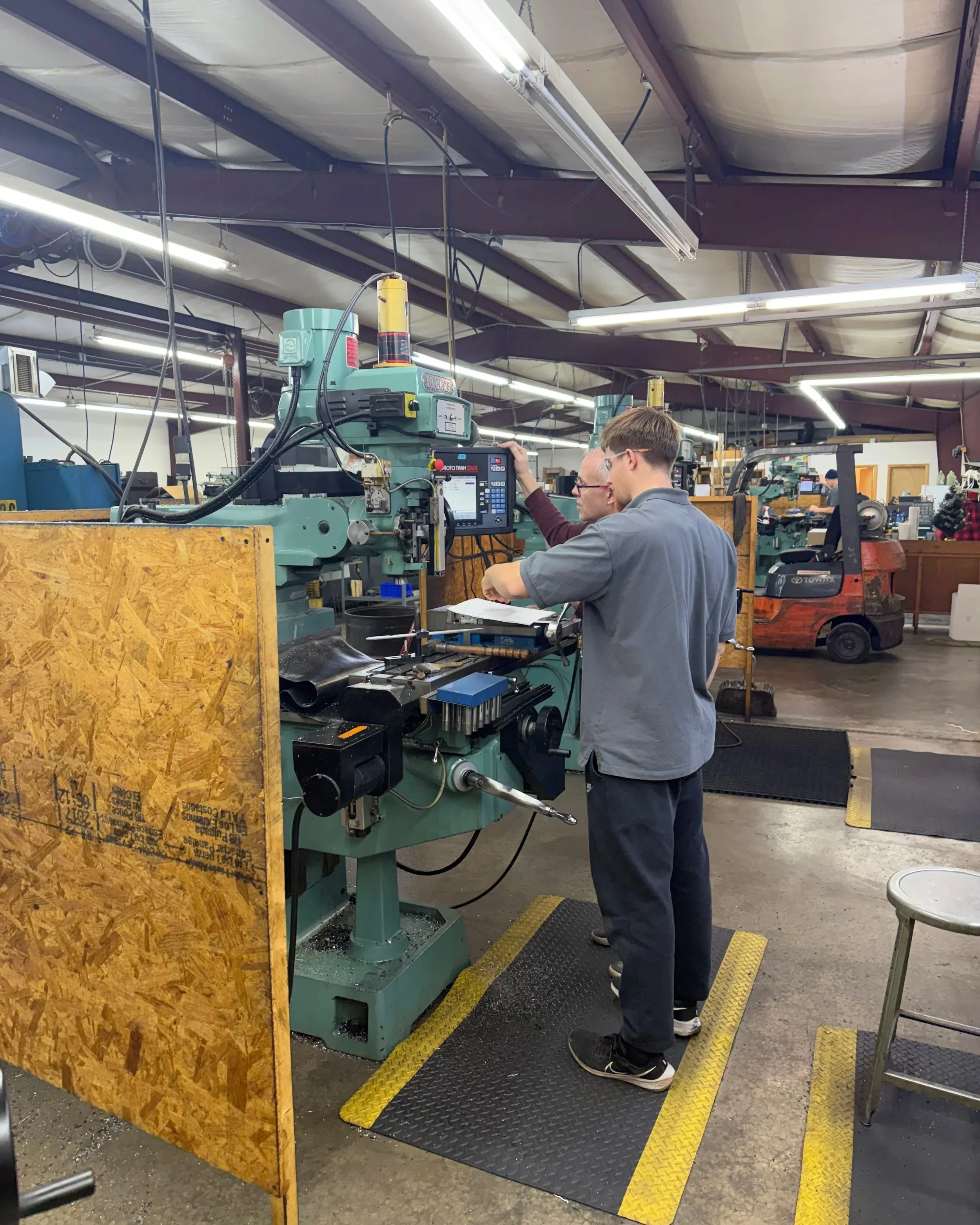 Two workers operate a large green industrial machine in a factory. There are fluorescent lights on the ceiling and various machinery in the background.