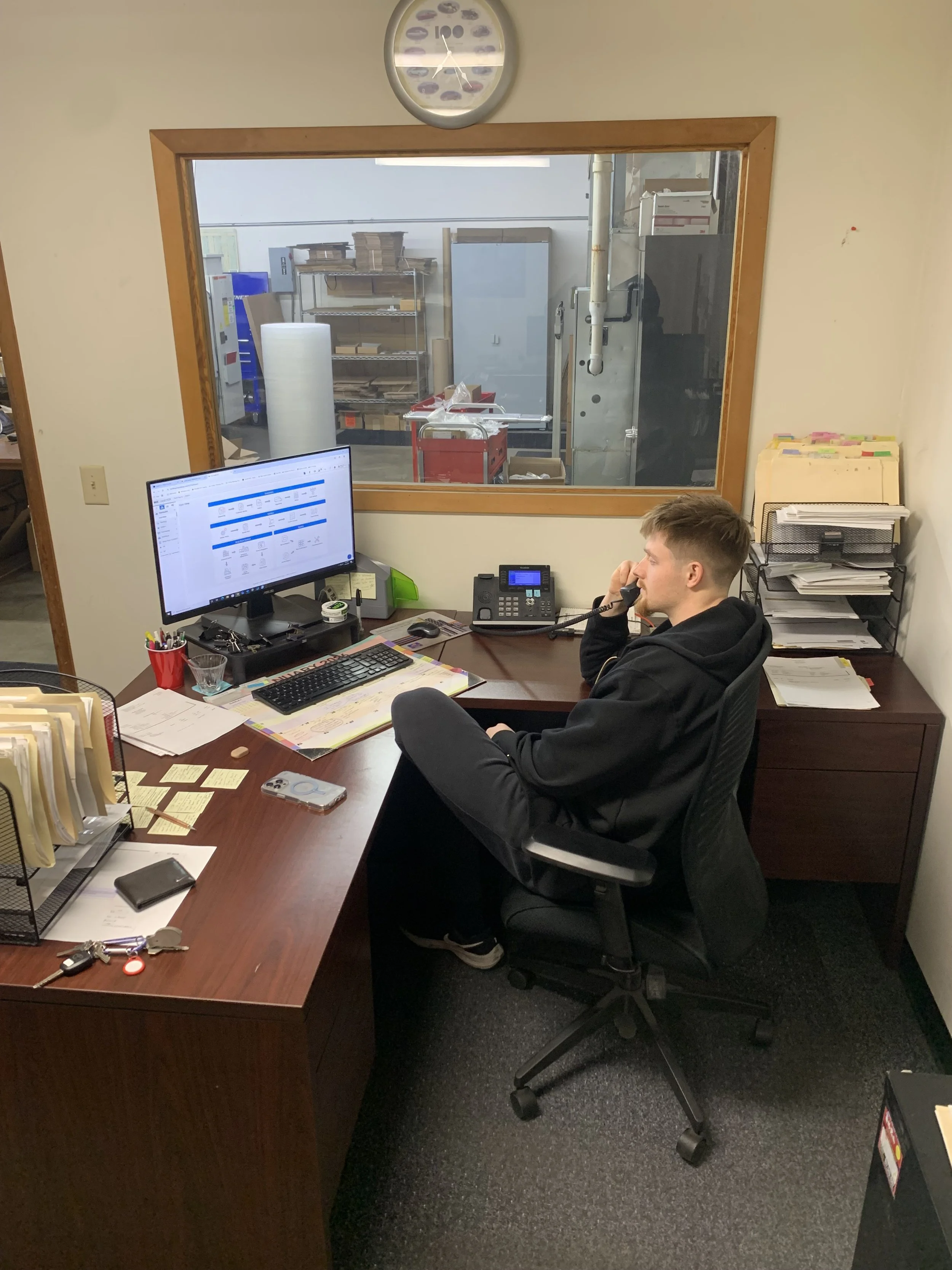 A man seated at a cluttered office desk talking on the phone, with a computer monitor, keyboard, and various papers and office supplies on the desk. A large window behind him shows an industrial kitchen or storage area.