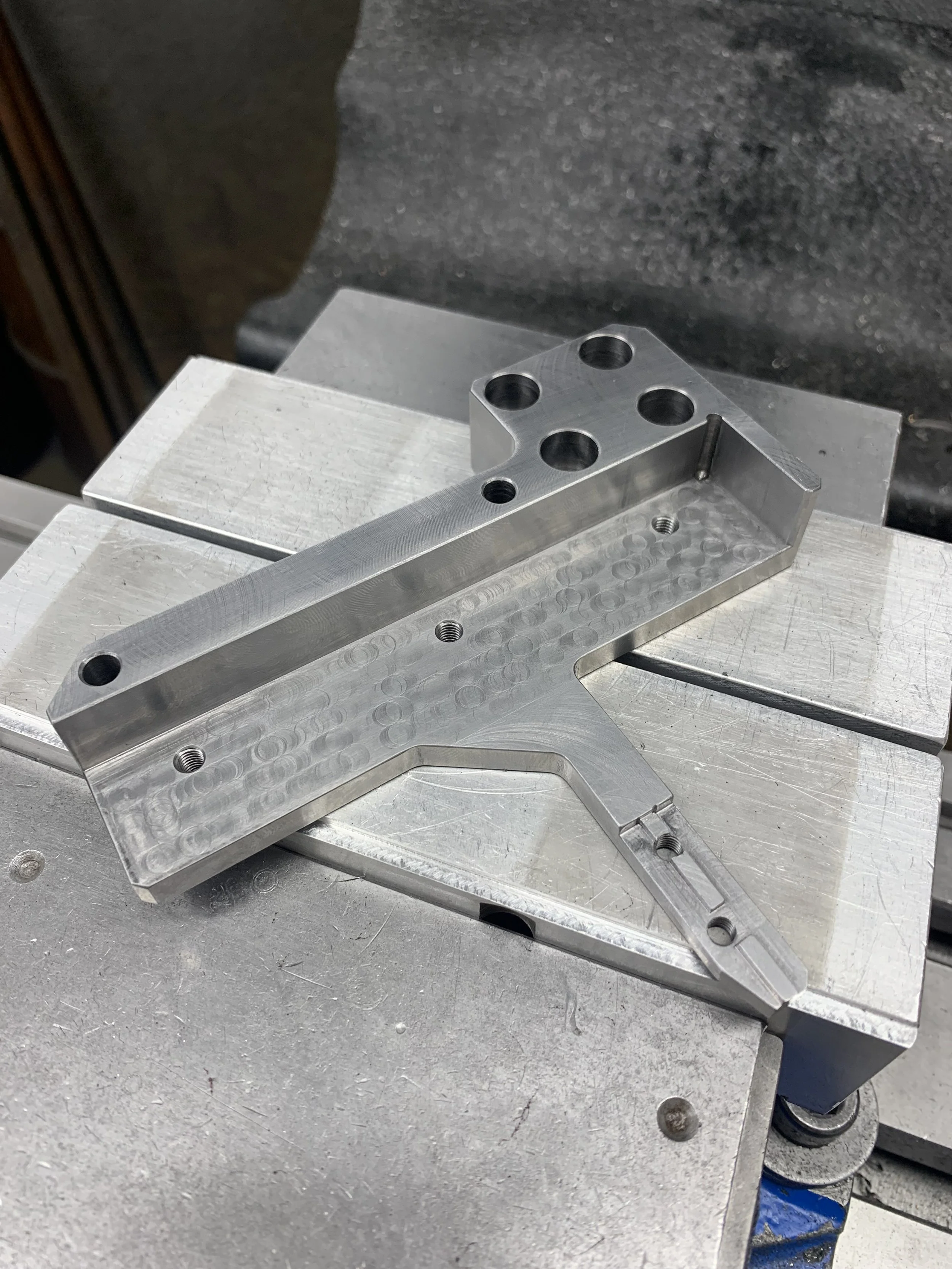 A machined metal component with drilled holes, placed on a metal worktable in a workshop.