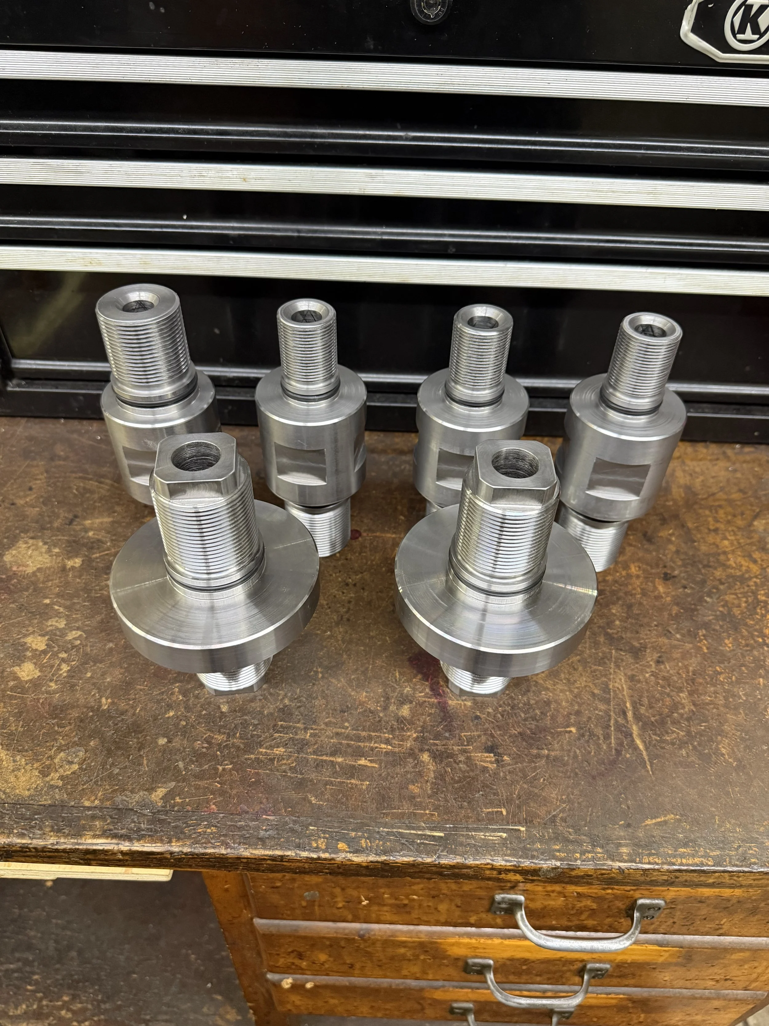 Six machined metal parts with threaded shafts placed on a wooden workbench in front of a black tool chest.