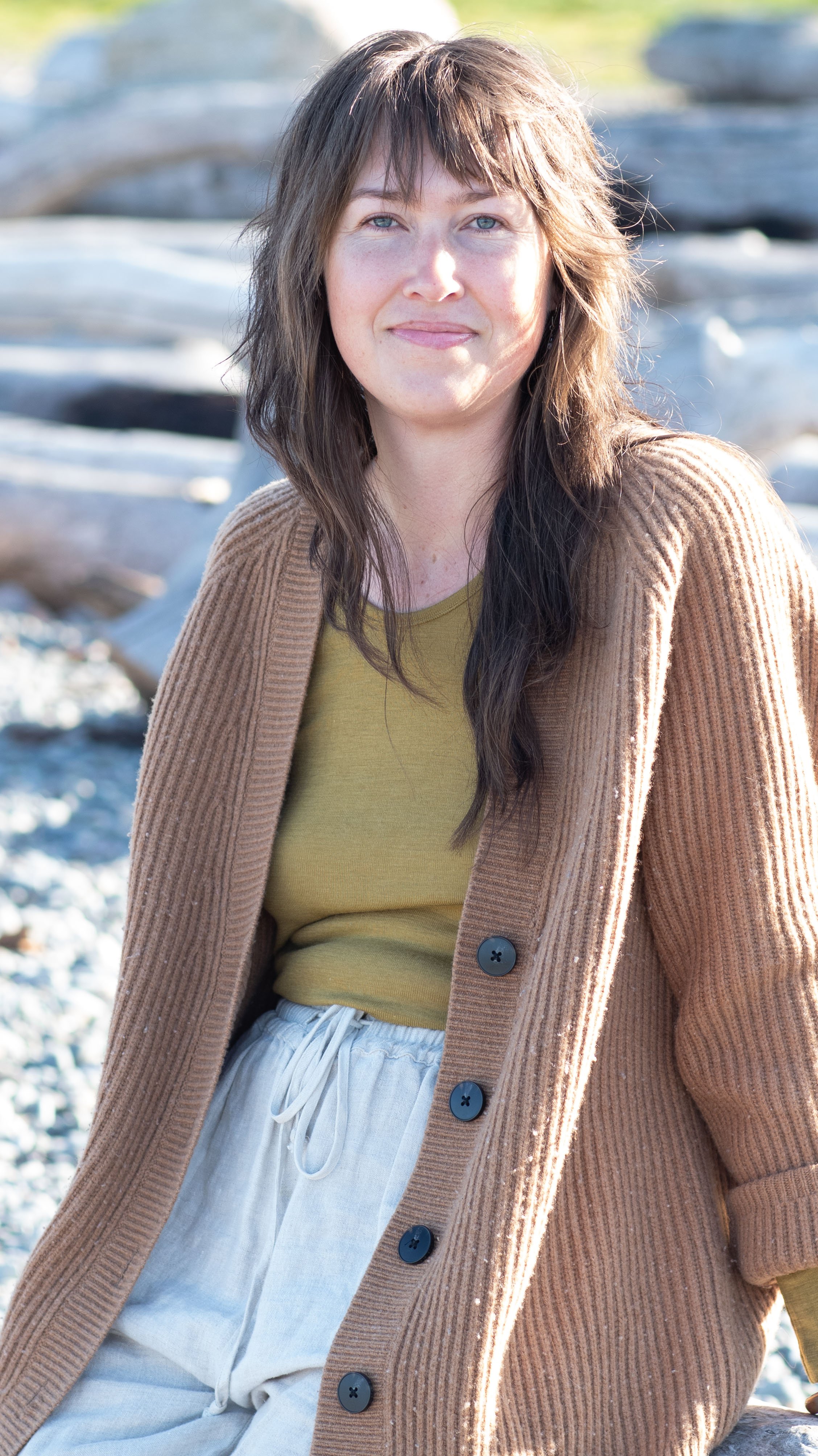 A woman with brown hair and blue eyes outdoors sitting on rocks near water, wearing a brown cardigan over a yellow shirt and white pants.