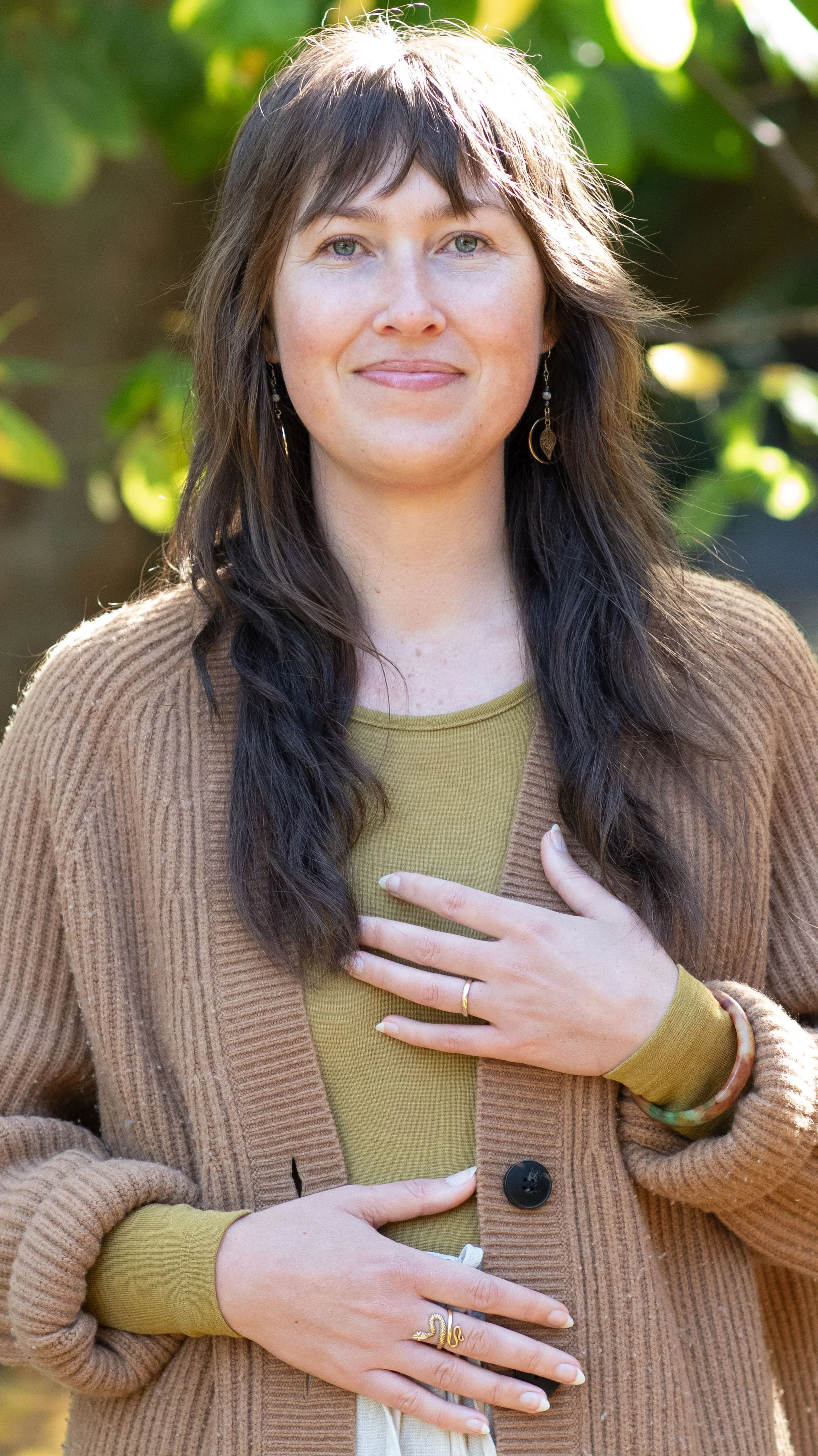 A woman with long brown hair and blue eyes smiling outdoors, wearing a brown cardigan over a green shirt, with her left hand on her chest and right hand on her stomach, in front of green leafy background.