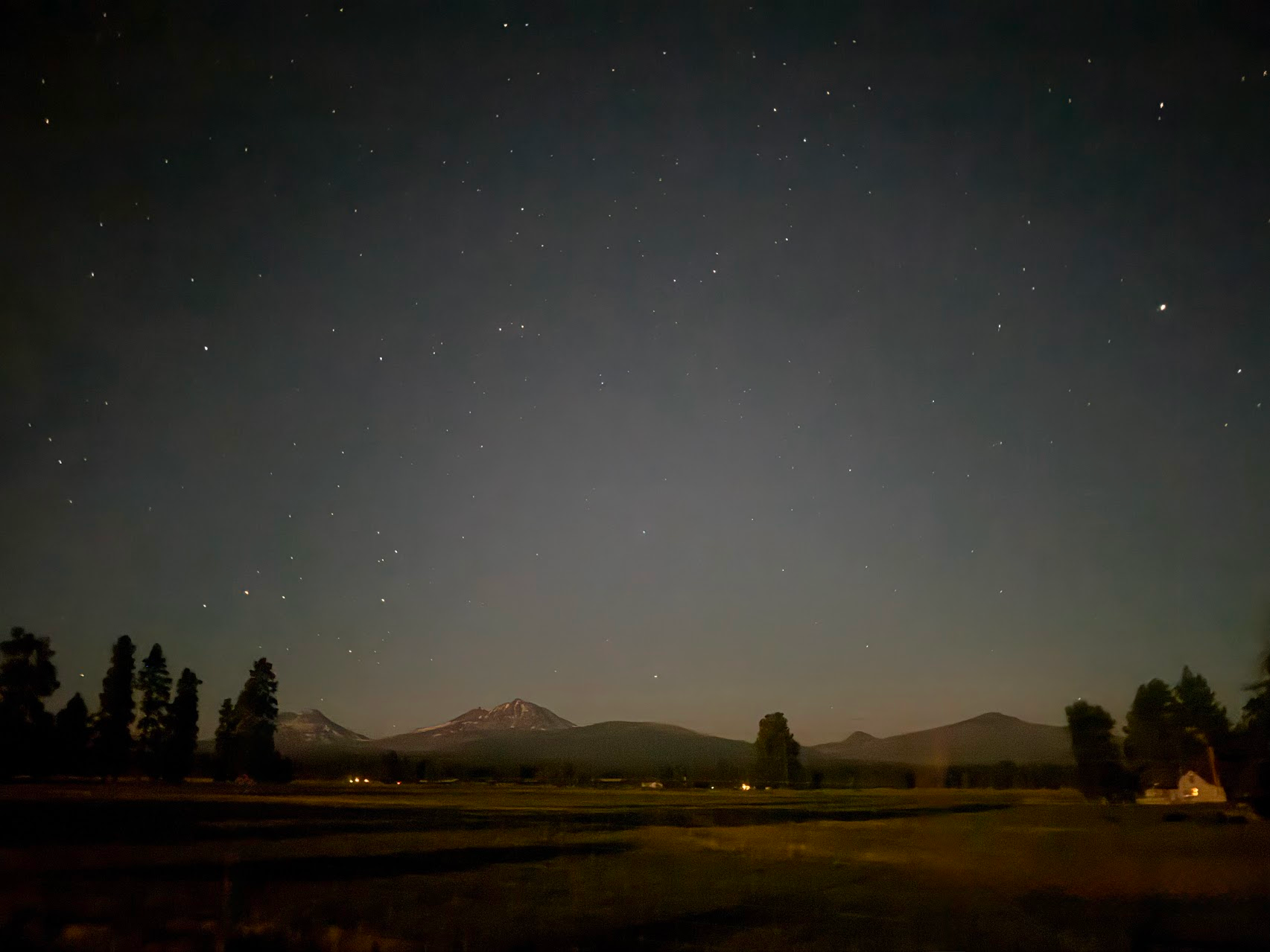 Night sky filled with stars over a landscape with trees and mountains in the distance.