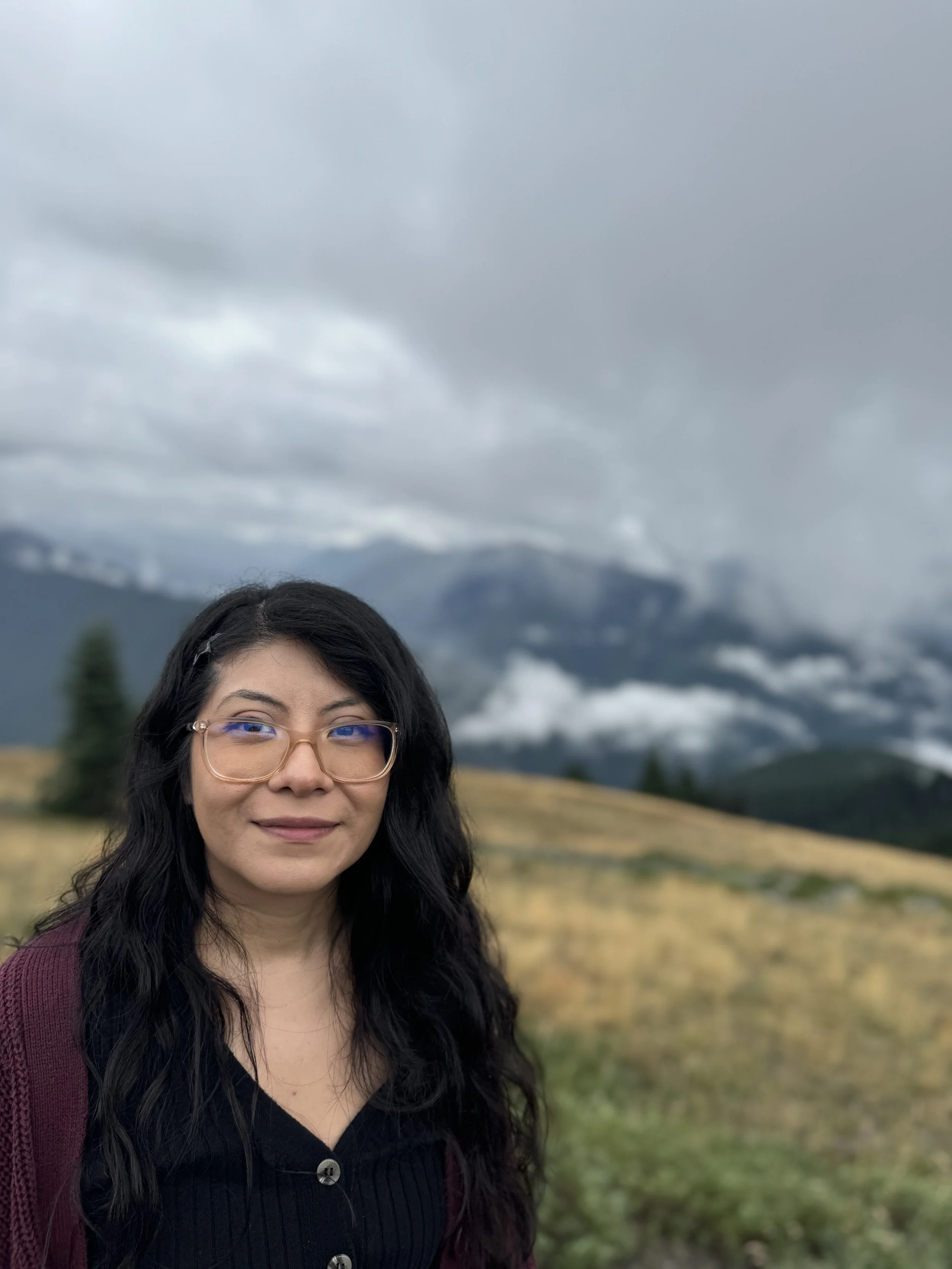 A woman with long black hair, glasses, and a black top standing outdoors in a mountainous landscape under cloudy skies.
