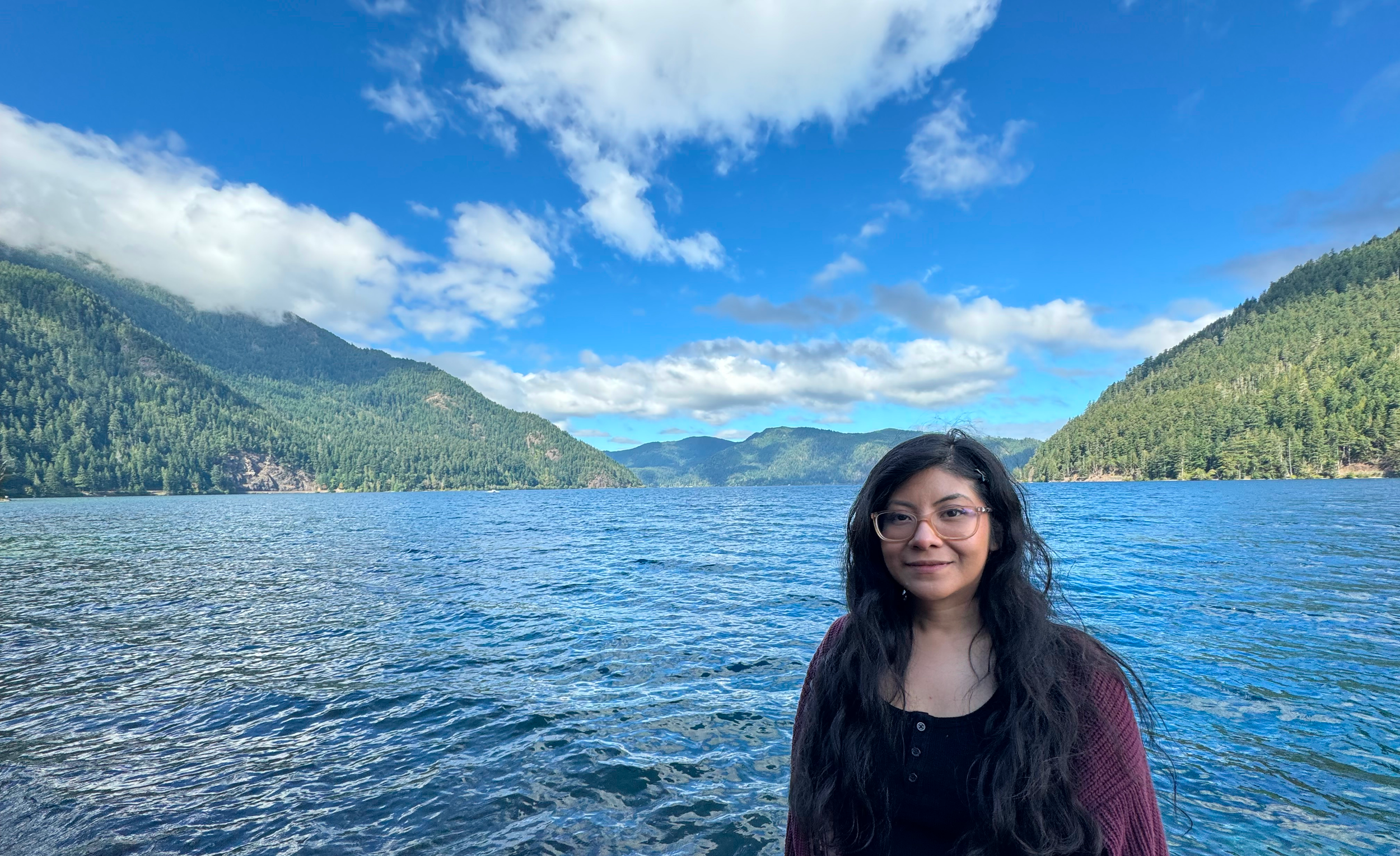 A woman with long dark hair, glasses, and a maroon cardigan standing near a large body of water surrounded by green mountains, under a partly cloudy blue sky.