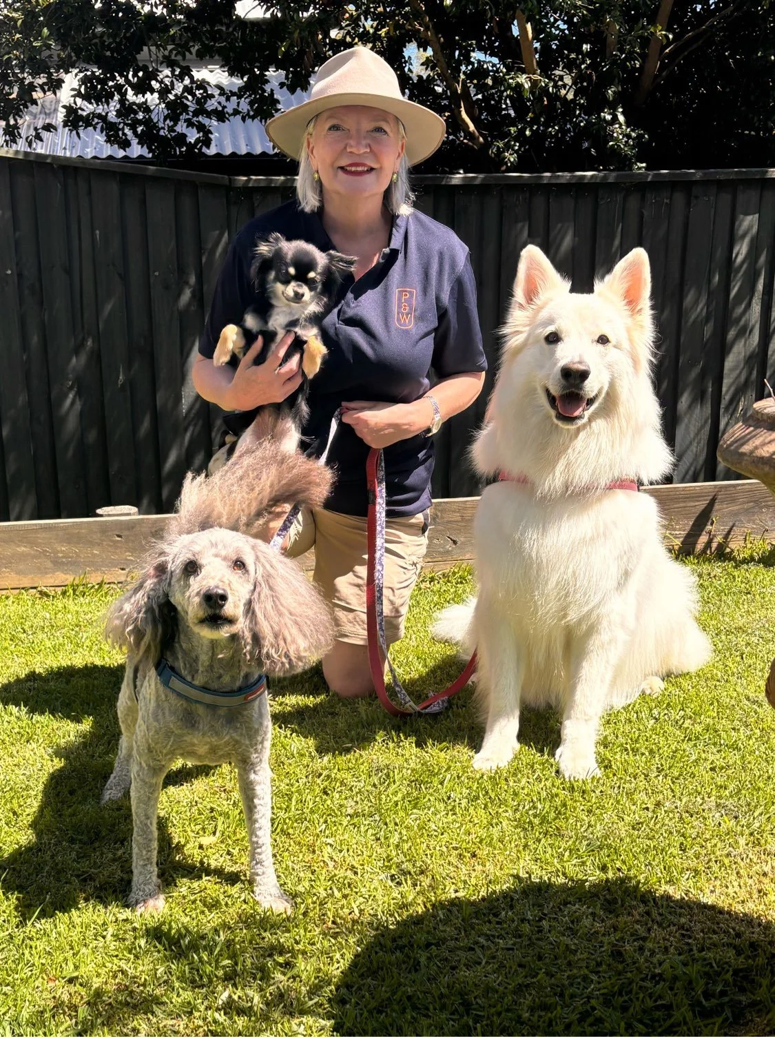 A woman wearing a beige hat and navy shirt kneels on a grassy lawn, holding a small black and tan dog. The woman is surrounded by three larger dogs: a cream-colored dog with fluffy fur sitting on the grass, a smaller curly-haired dog standing with a blue collar, and a black and tan dog in the woman's arm. Behind them, a black wooden fence and trees are visible.