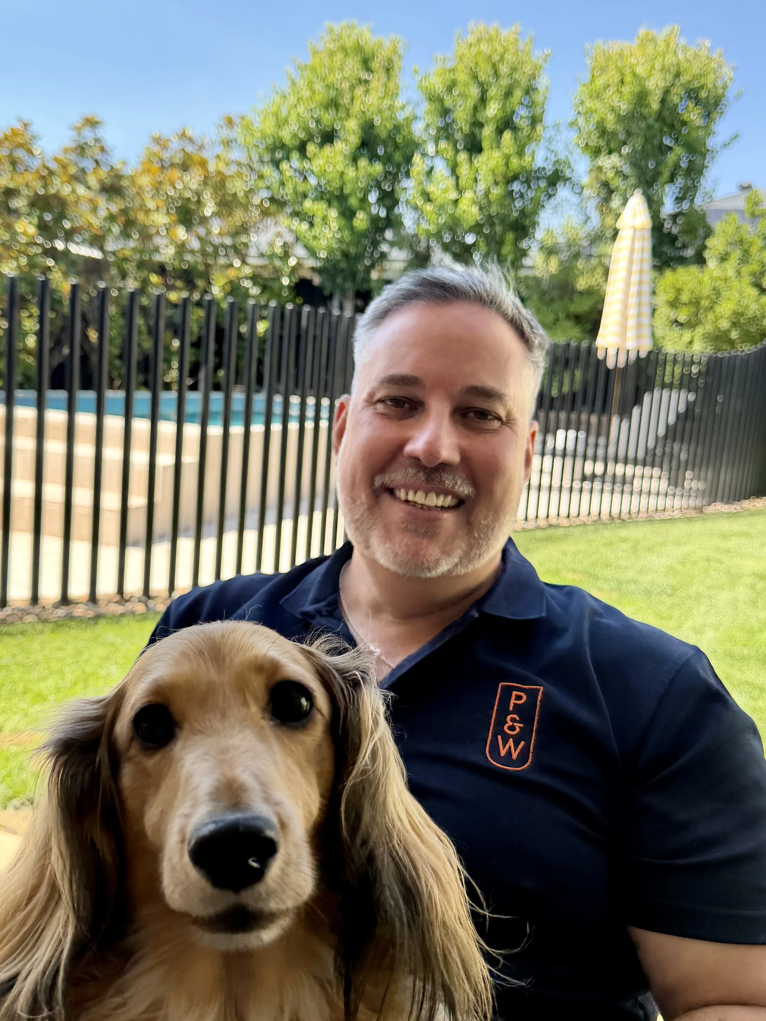 Smiling man taking a selfie with a golden retriever outdoors next to a pool, with green trees in the background.