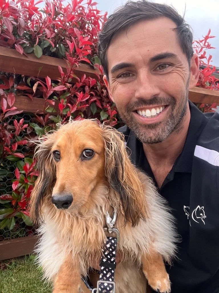 A man smiling and a golden retriever sitting outdoors with a background of red and pink foliage.