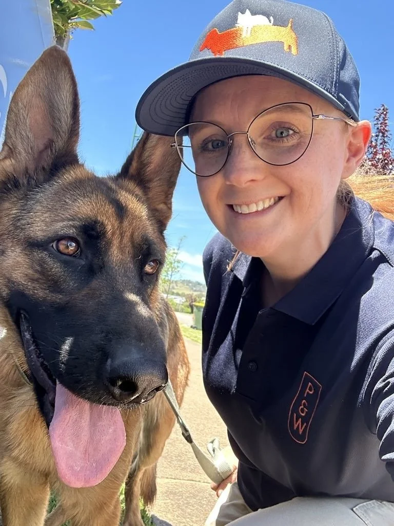 A woman smiling and taking a selfie with a large German Shepherd dog outdoors on a sunny day, with trees and a blue sky in the background.
