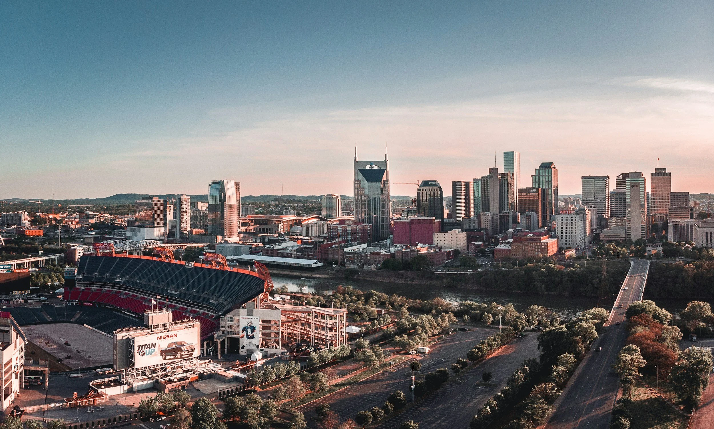 A city skyline during sunset with tall modern buildings, a stadium in the foreground, and a river flowing through the city, surrounded by roads and trees.