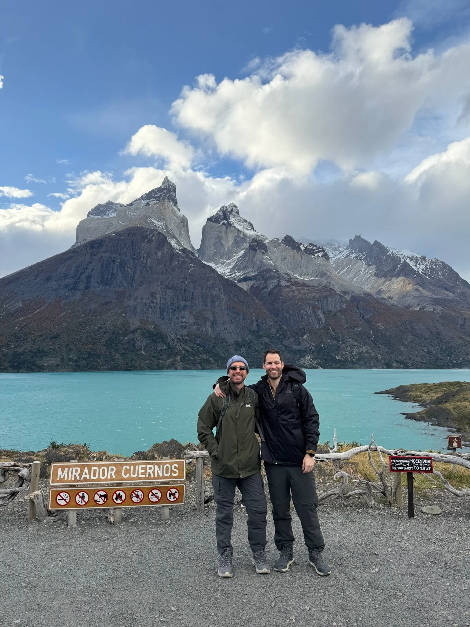 Two men stand in front of a scenic mountain lake with rugged snow-capped peaks in the background. They are smiling and wearing outdoor jackets and backpacks. The area is fenced with signs, including one that says 'Mirador Cuernos' and another warning in Spanish not to touch or feed wildlife.