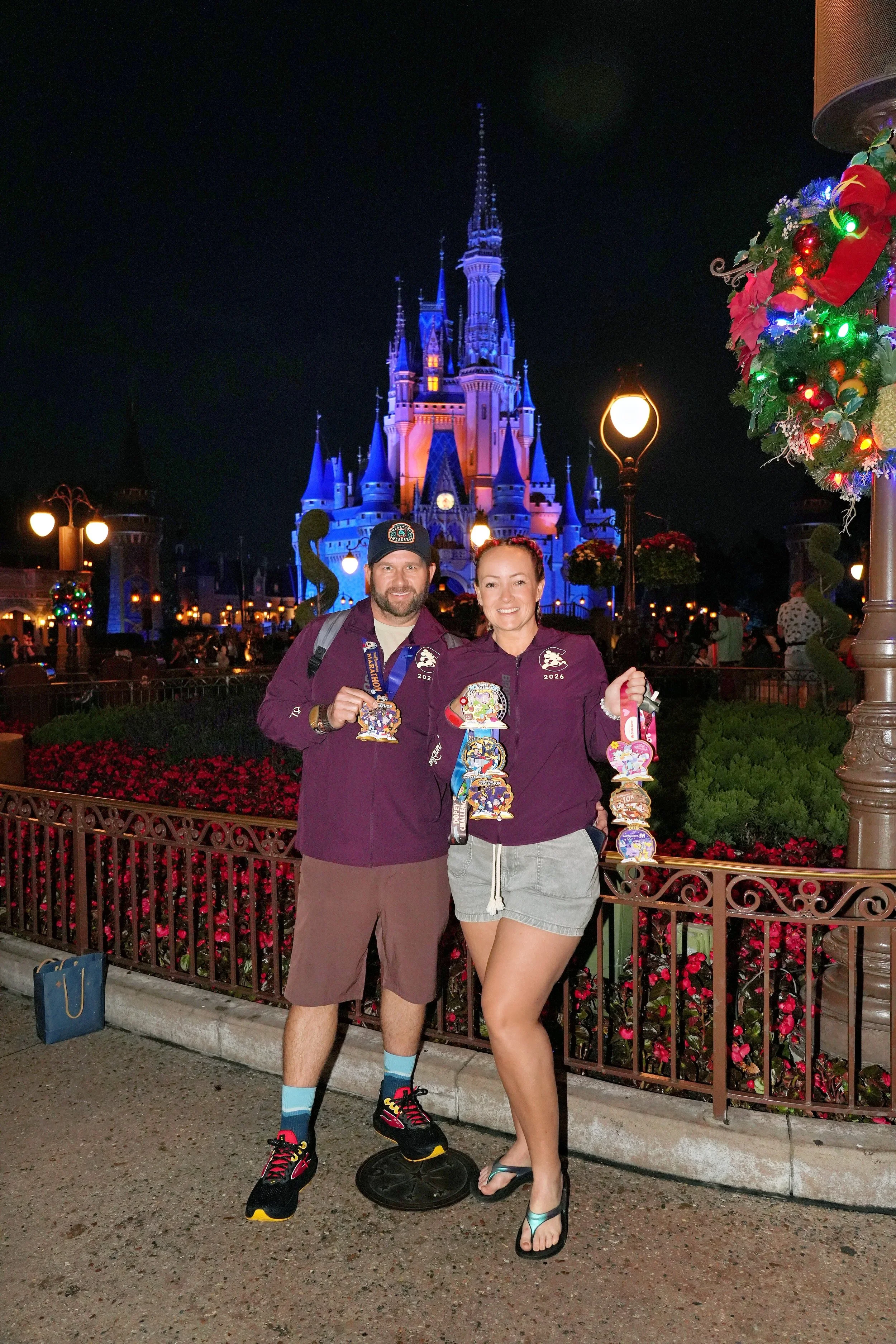 A man and woman standing in front of Cinderella Castle at night, holding Disney medals, with Christmas decorations and crowd in the background.