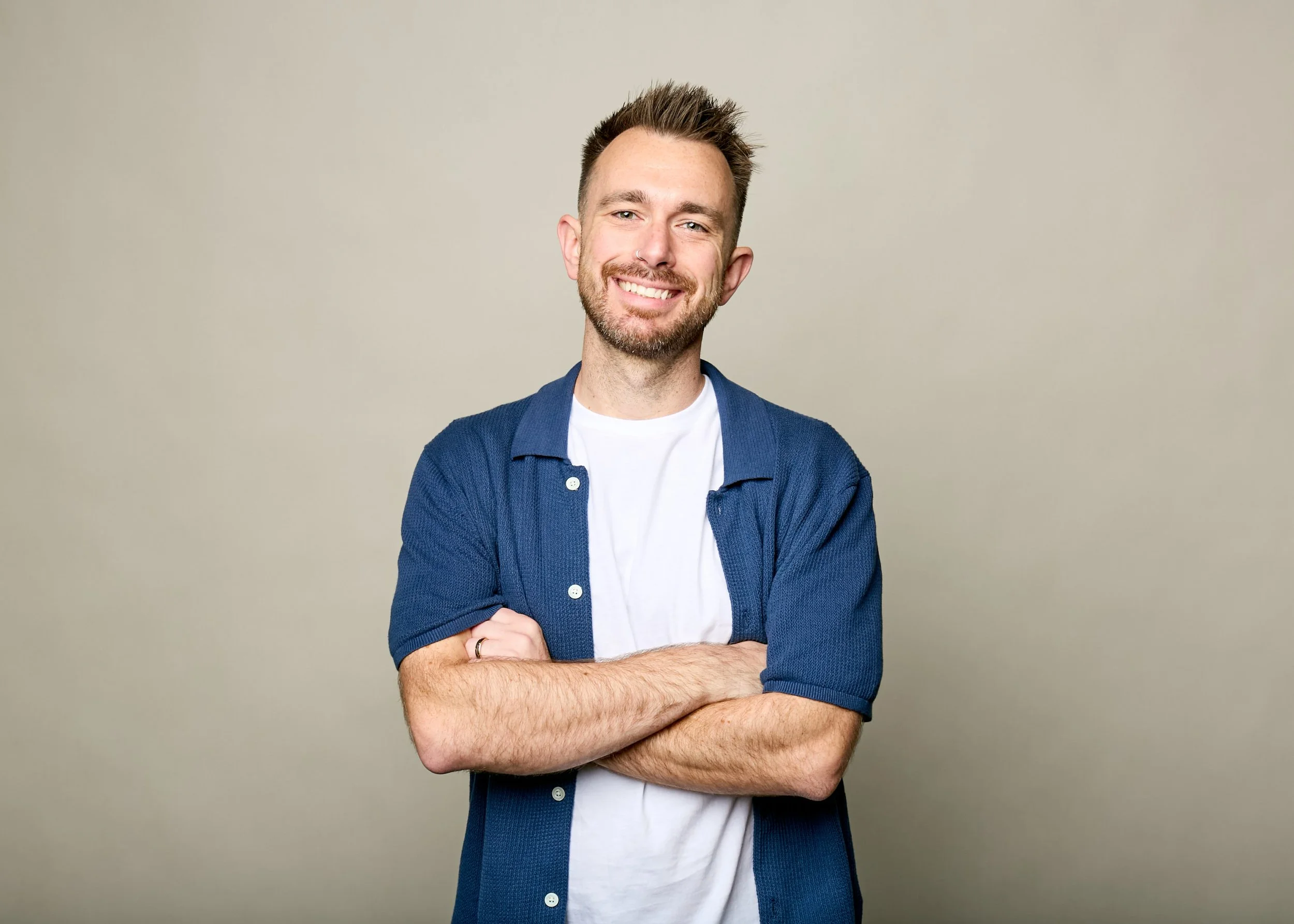 A smiling man with short brown hair, a beard, and a nose piercing, wearing a white t-shirt and a blue shirt, standing with arms crossed against a plain, light-colored background.
