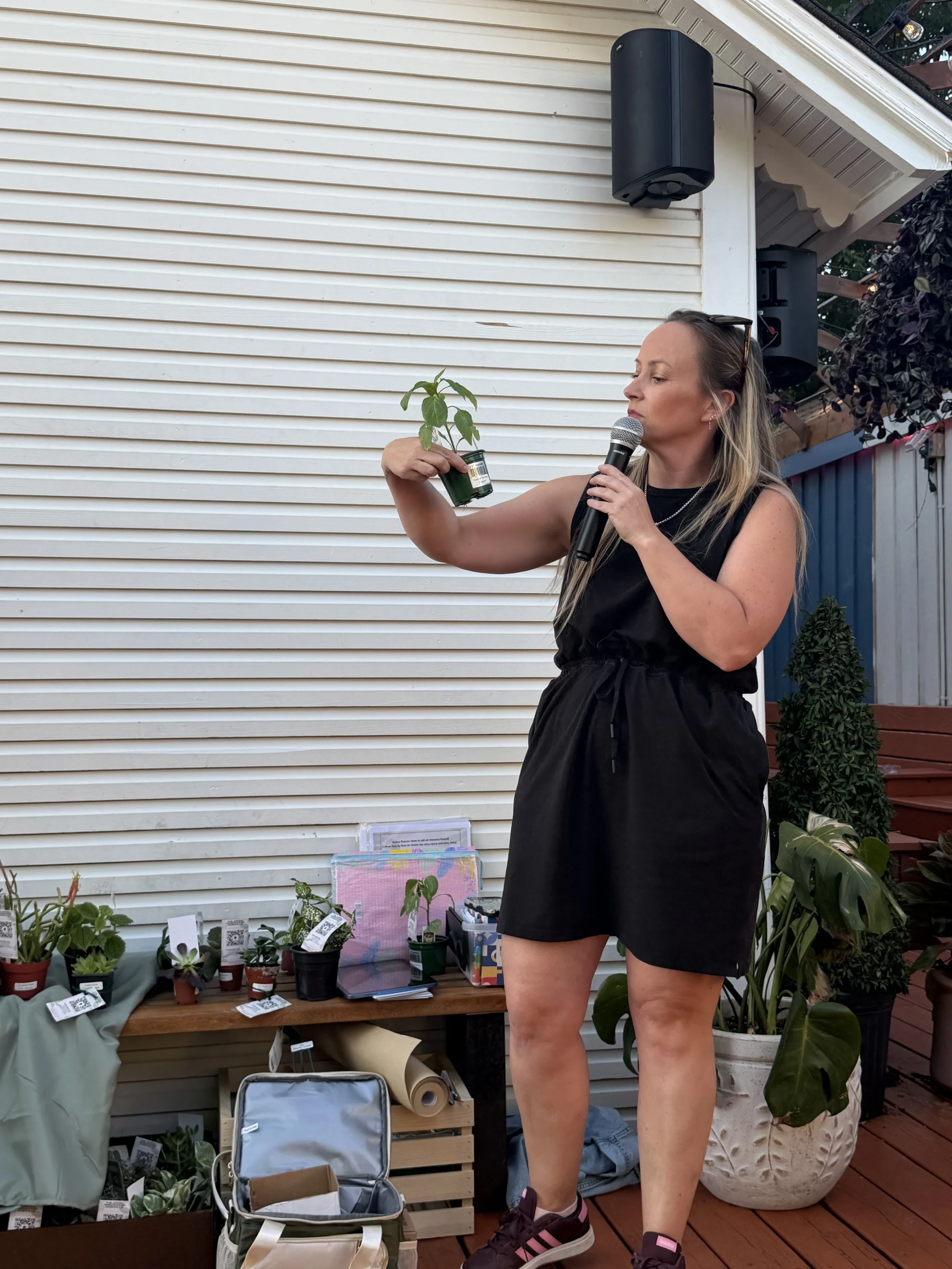 A woman with long blonde hair, wearing a black sleeveless dress and sneakers, stands outdoors on a wooden deck with a microphone in her right hand. She is holding a small potted plant in her left hand and appears to be speaking or presenting. There are various potted plants and gardening supplies on a table and shelves behind her, with a white horizontal siding house wall in the background.