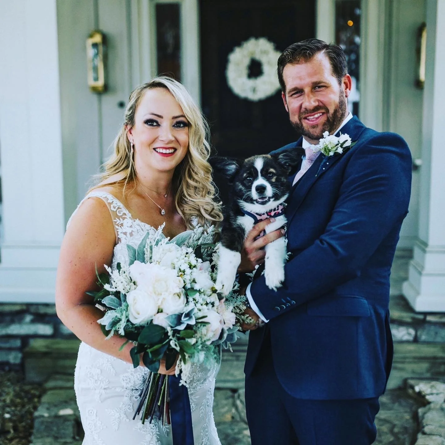A couple in wedding attire holding a small black and white dog with a pink collar, indoors in front of a dark doorway with a floral wreath, celebrating their wedding.
