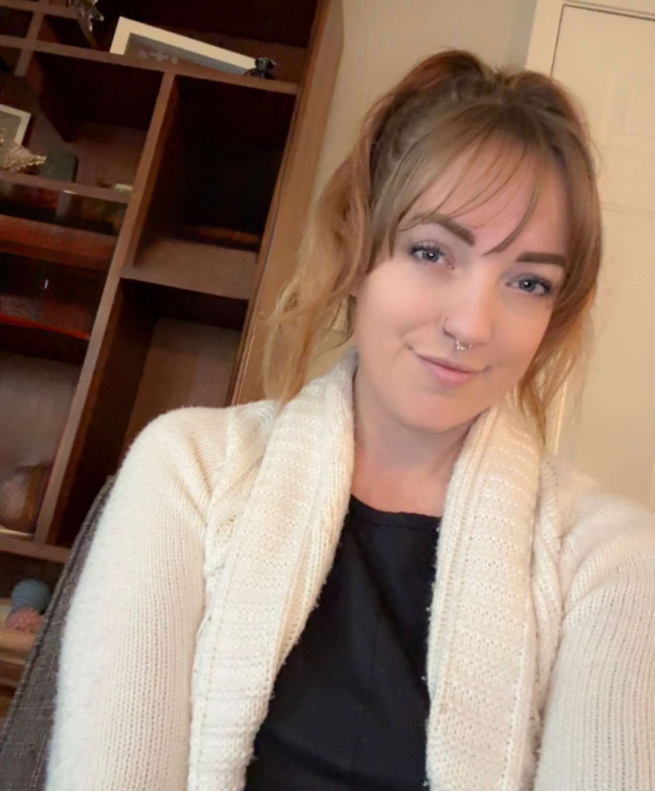A woman with light brown hair and a septum piercing, wearing a cream-colored sweater and black shirt, smiling in a room with a wooden bookshelf and beige wall in the background.
