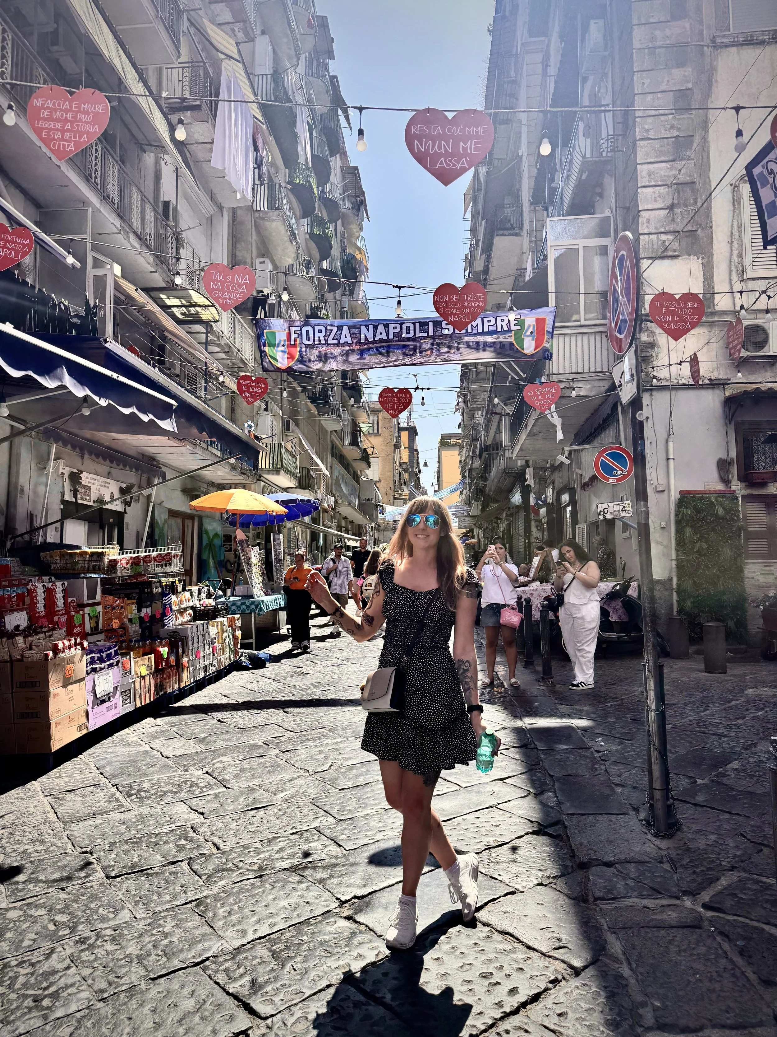 A woman with sunglasses, a black polka dot dress, and white sneakers walking on a cobblestone street in a busy city, carrying a green bottle. There are street vendors, people walking, and buildings with balconies, and hanging decorations in the background.