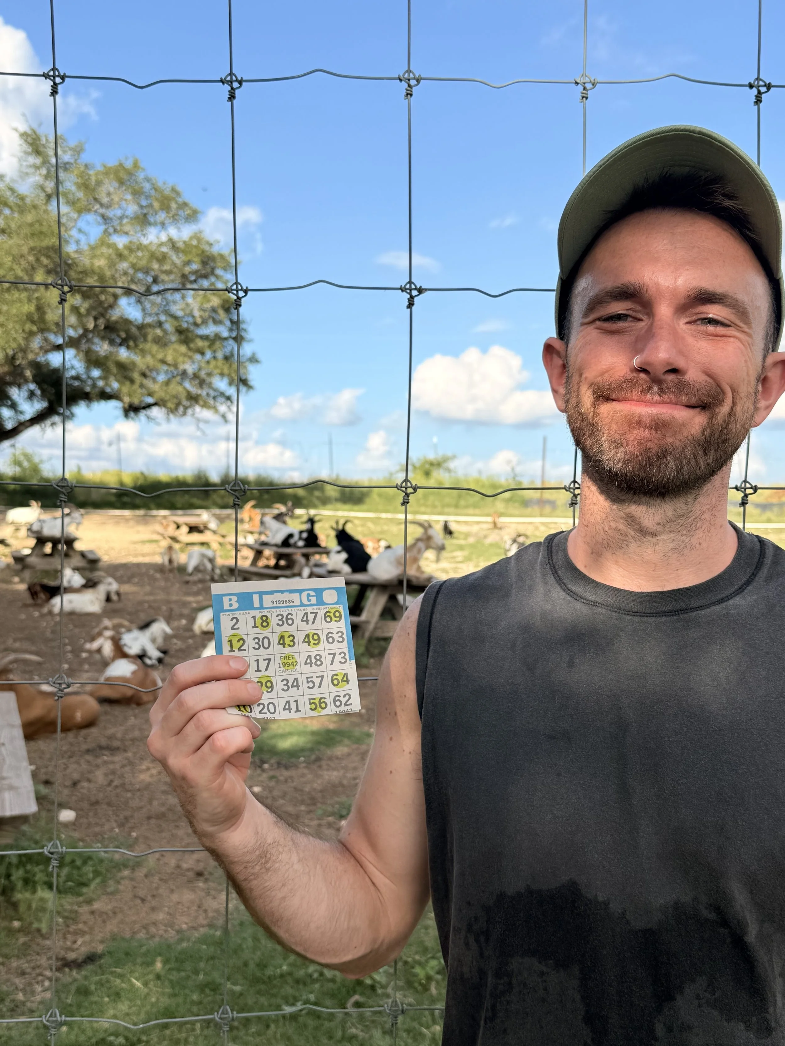 A man with a beard, wearing a green cap and sleeveless shirt, smiling and holding a lottery ticket in front of a wire fence at a farm with goats and trees in the background.