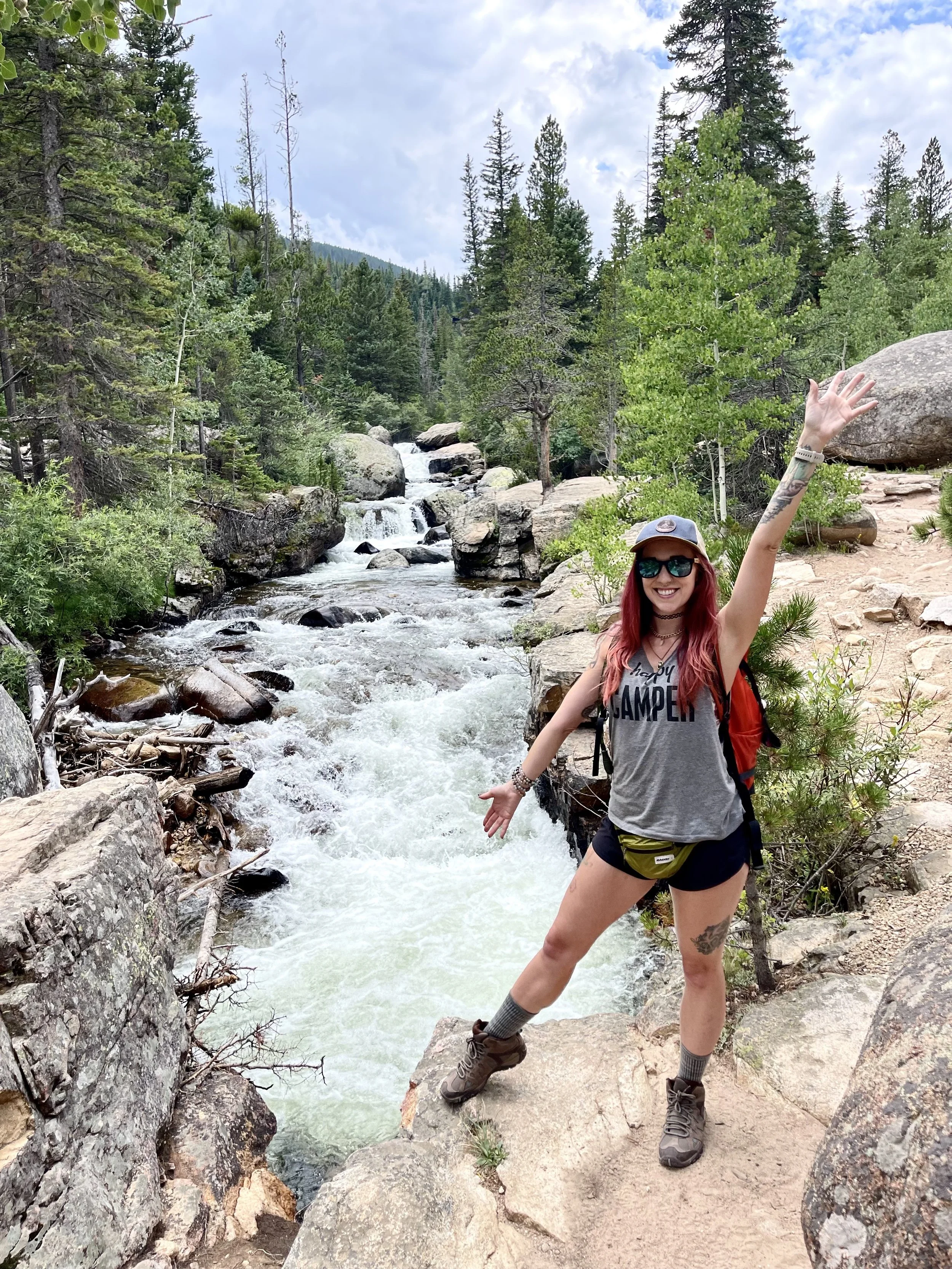 A woman with red hair, sunglasses, a gray t-shirt, shorts, hiking boots, and a cap, smiling and posing with arms raised, standing on a rocky trail next to rushing river in a forested mountain landscape with trees and clouds in the sky.