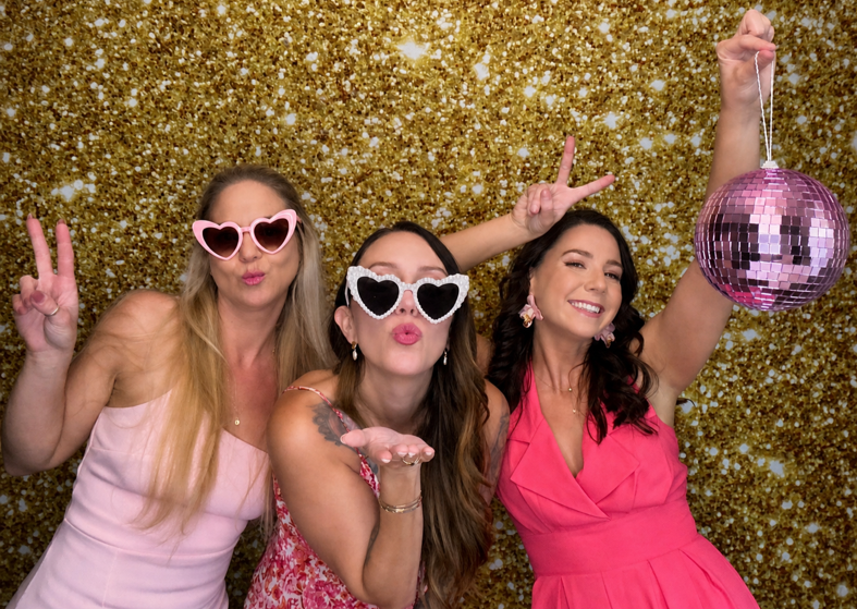 Women posing at a Photo Booth with heart shaped sunglasses and a gold backdrop at a wedding in Winter Springs, FL, creating fun and memorable moments.