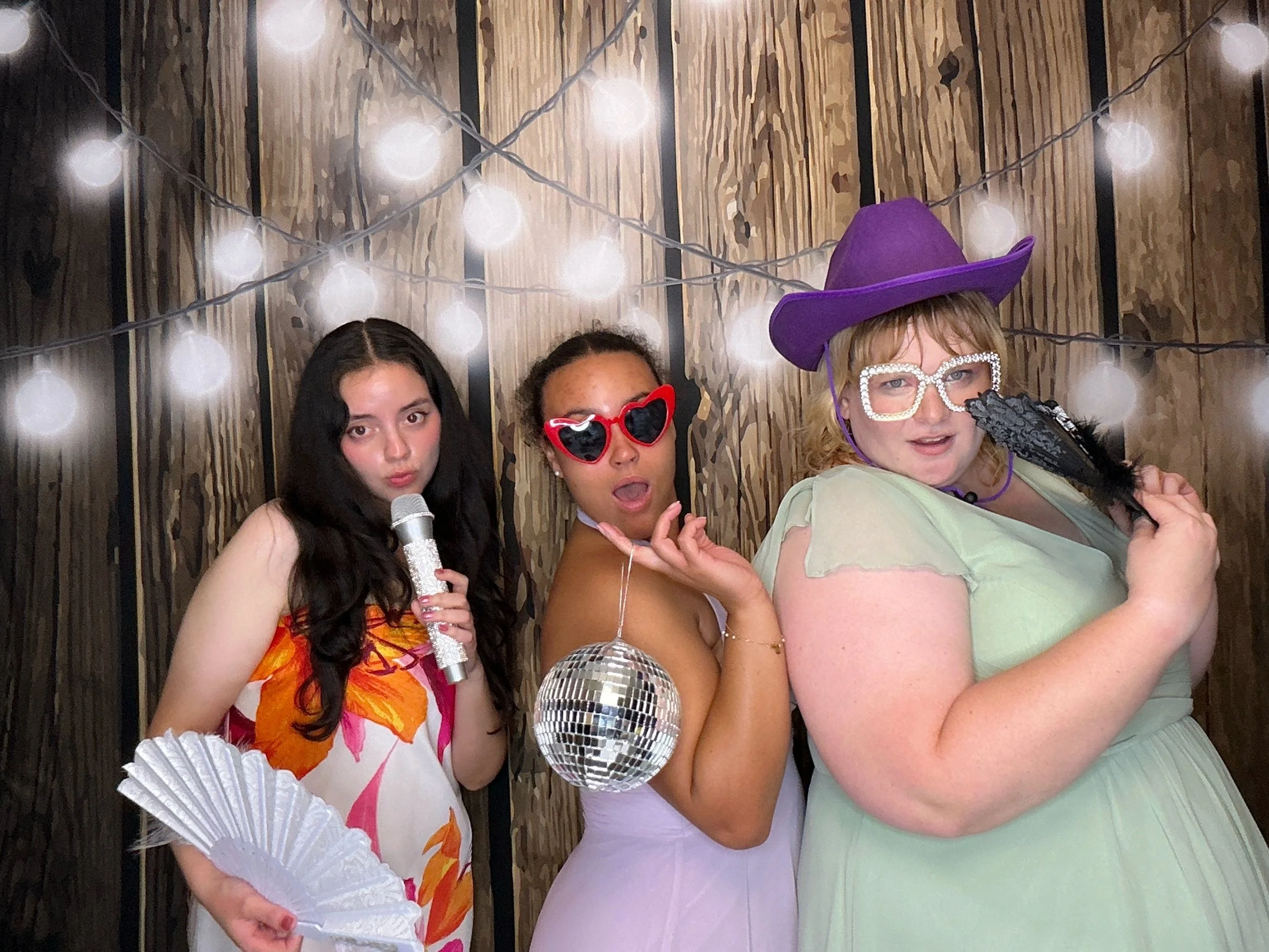 Three guests posing with fun Photo Booth props in front of a wood backdrop with string lights at a wedding reception in Oviedo, Florida.