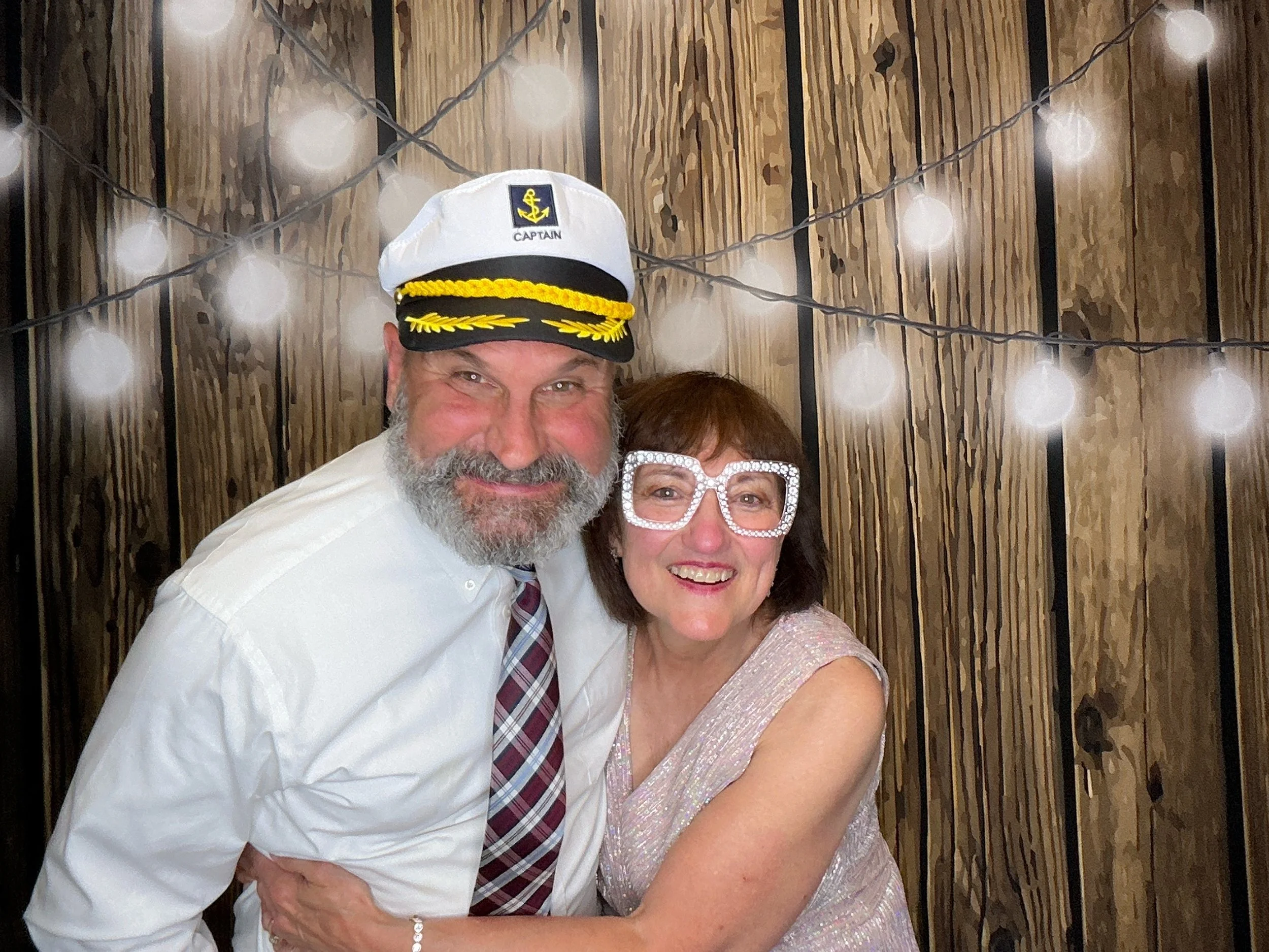 Couple smiling with fun Photo Booth props in front of a wood backdrop with string lights at a wedding reception in Winter Springs, Florida.