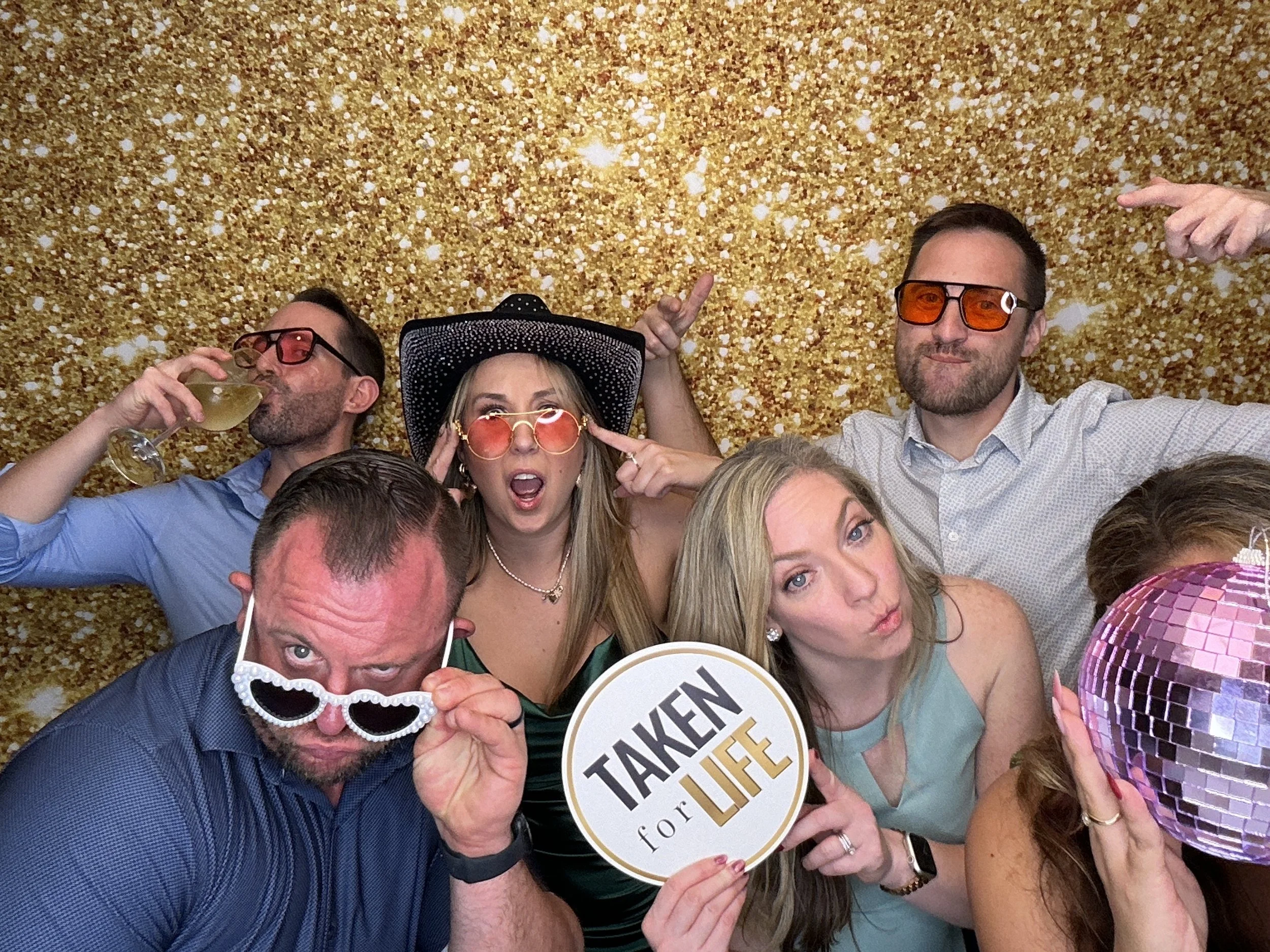 Guests posing with Photo Booth props in front of a gold sequin backdrop during a wedding celebration with Pop & Pose Events