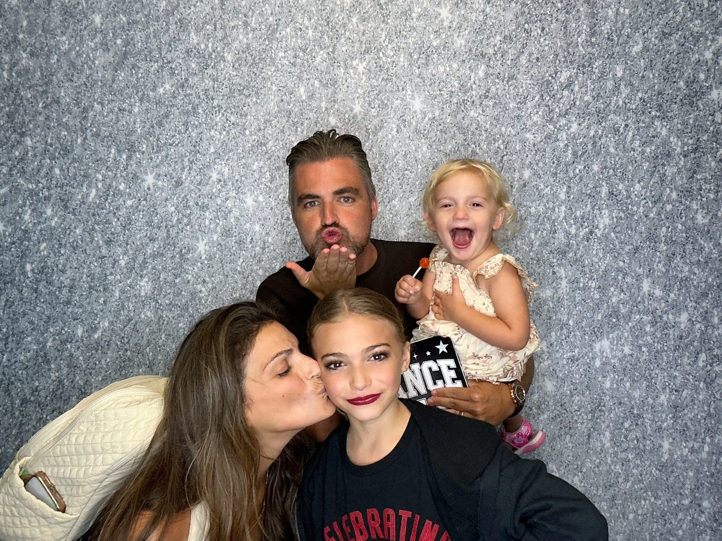 A man with slicked-back dark hair and a beard blowing a kiss, holding a young girl with blonde curly hair and a beige dress. They are surrounded by other children; one girl with long brown hair and dark lipstick, and another girl with light skin and light brown hair. The background is sparkly silver.