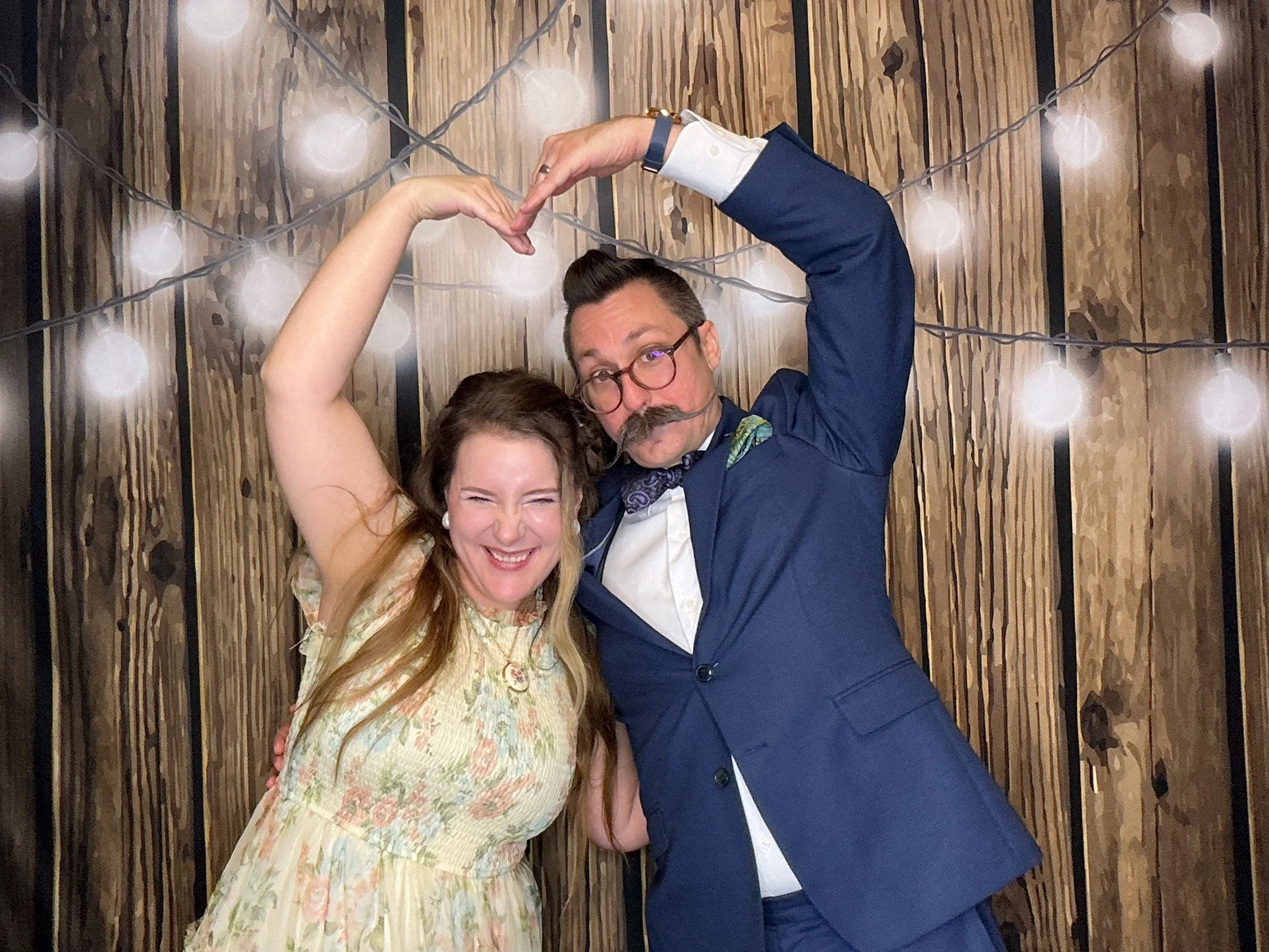 Couple making a heart shaper with their hands in the Photo Booth at a wedding reception in front of a wood backdrop with string lights in Oviedo, Florida.