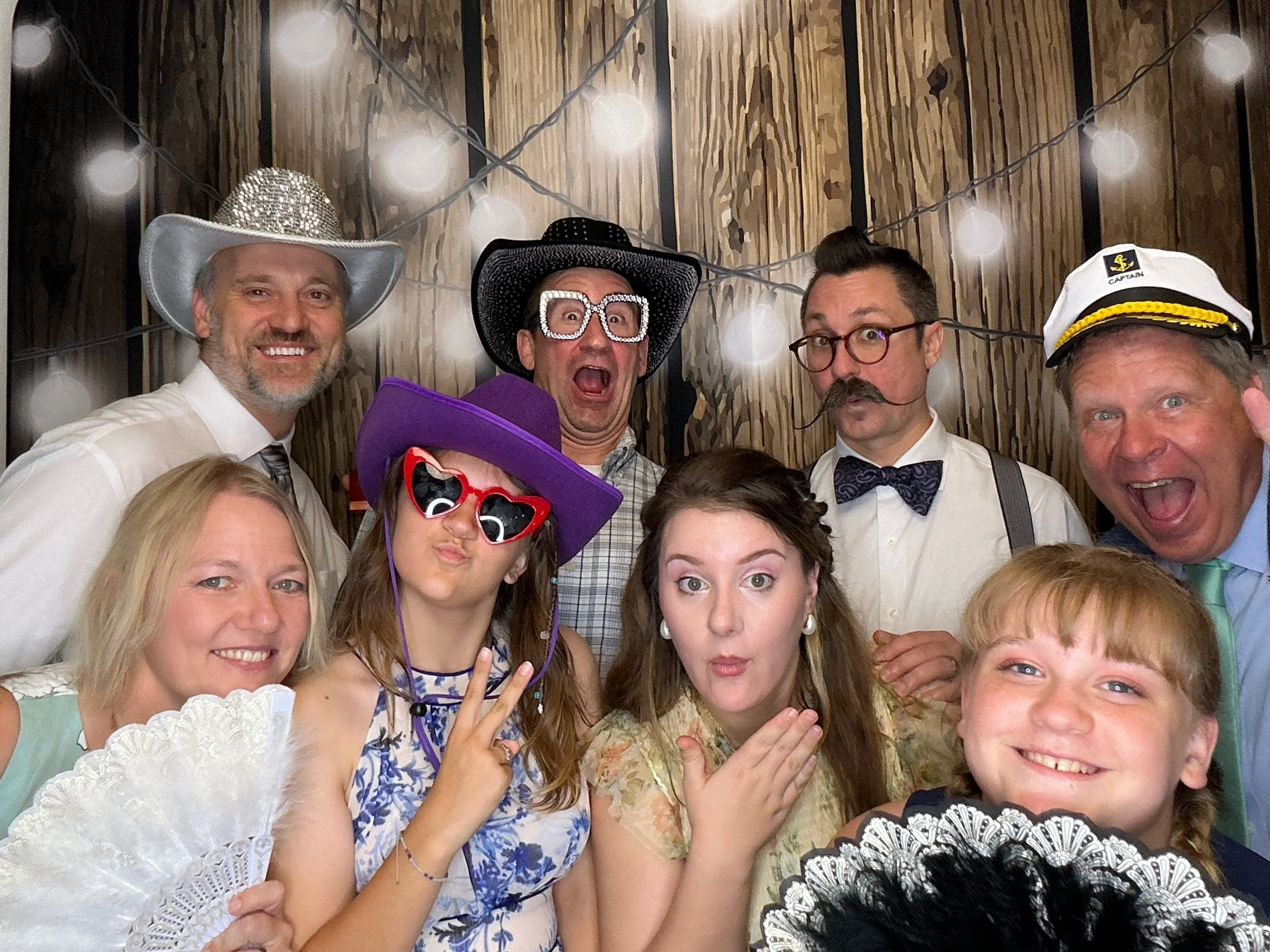 Guests posing with fun Photo Booth props in front of a wood backdrop with string lights at a wedding reception in Apopka, Florida.