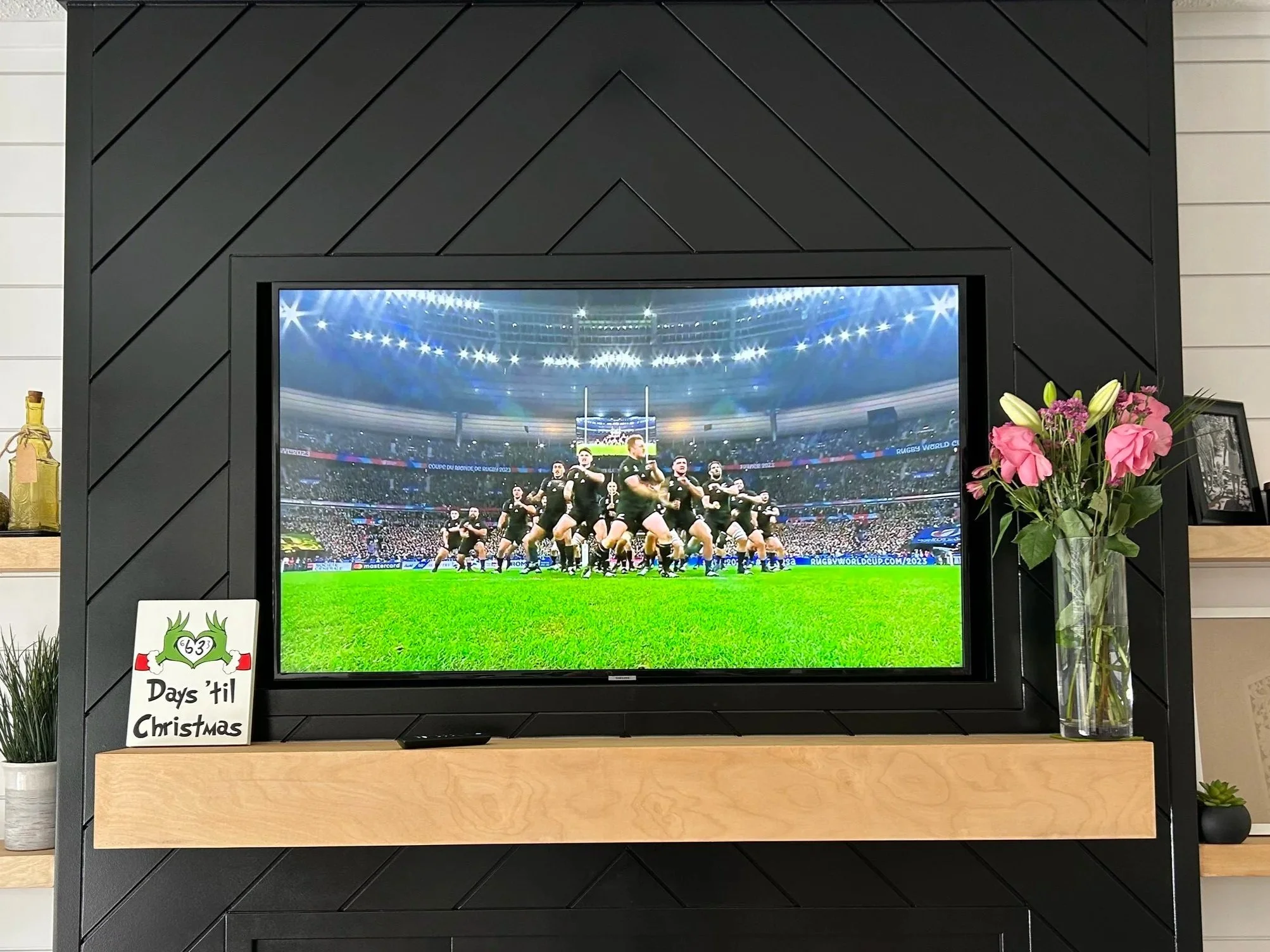 A television on a black geometric-patterned wall displaying a rugby game with players on the field. To the right of the TV, there is a vase with pink and white flowers on a wooden shelf. To the left, a small sign reads 'Days 'til Christmas' with a green dragon illustration. There are also small plants on the shelves.