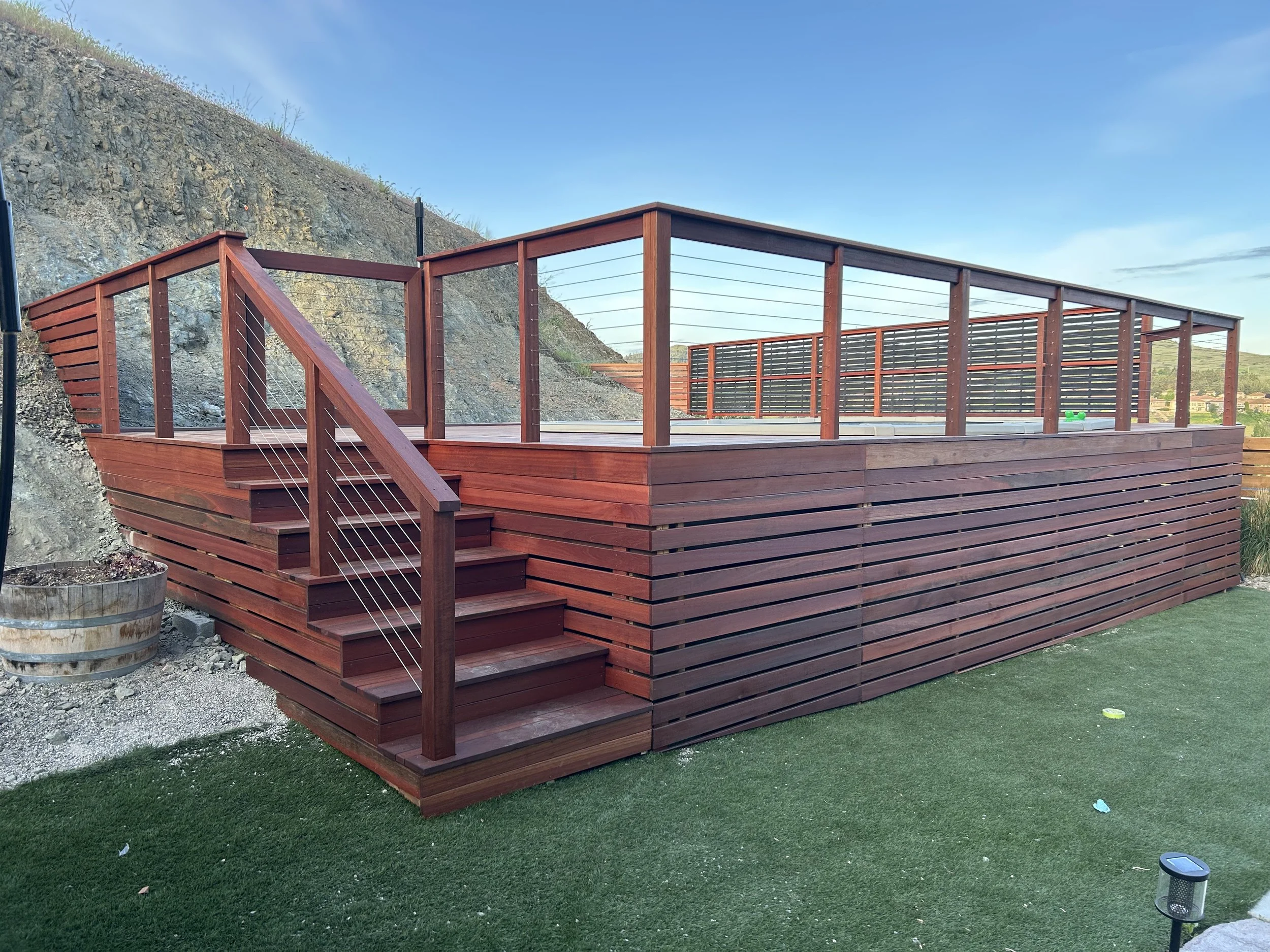 A large wooden deck with stairs, railings, and horizontal slats, built on a sloped terrain with a hillside on the left and a grassy yard in the front. The sky is blue with some scattered clouds.