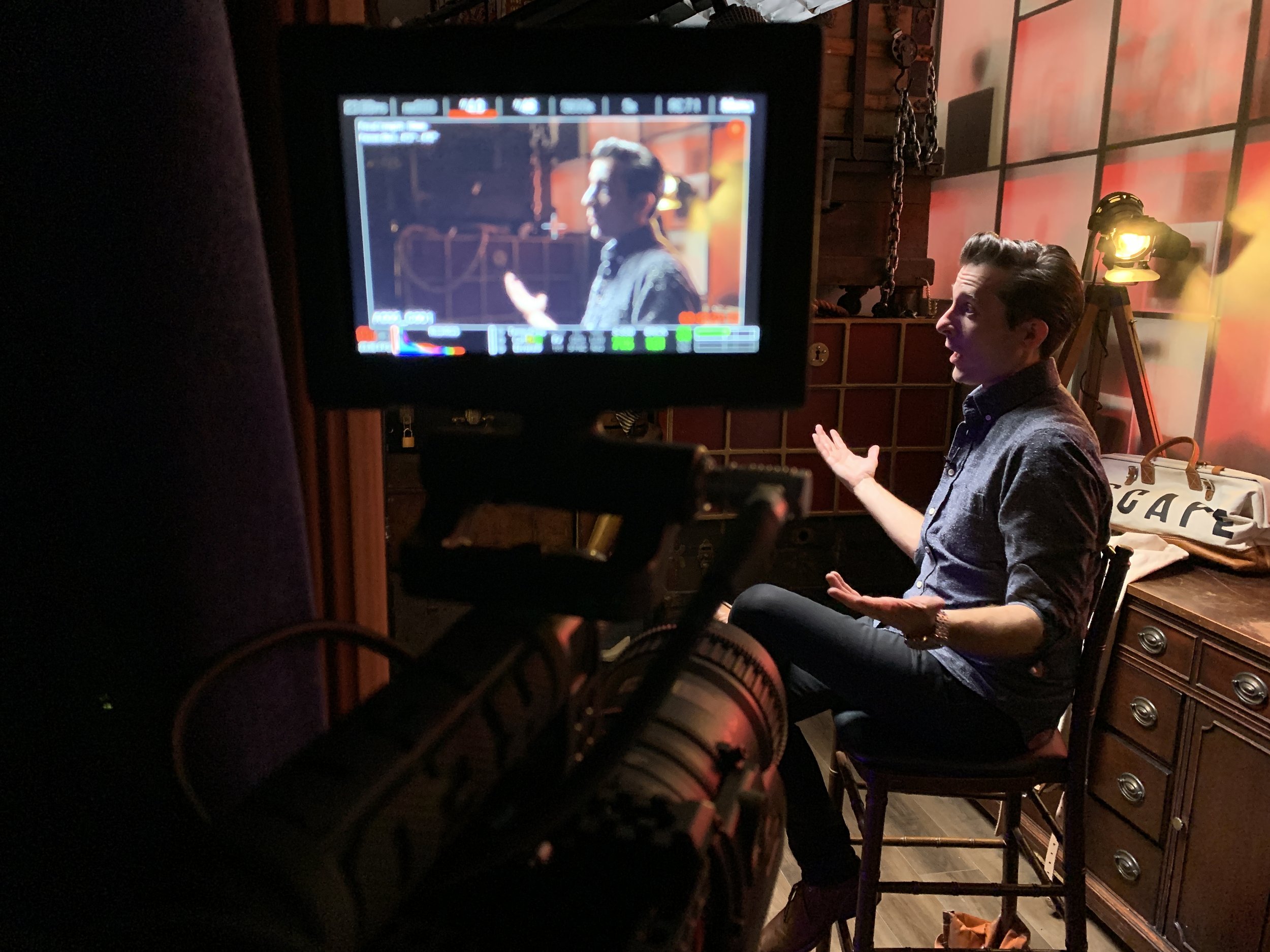 A person is conducting an interview in a studio with a warm background and studio equipment. The person's facial expression suggests they are speaking.