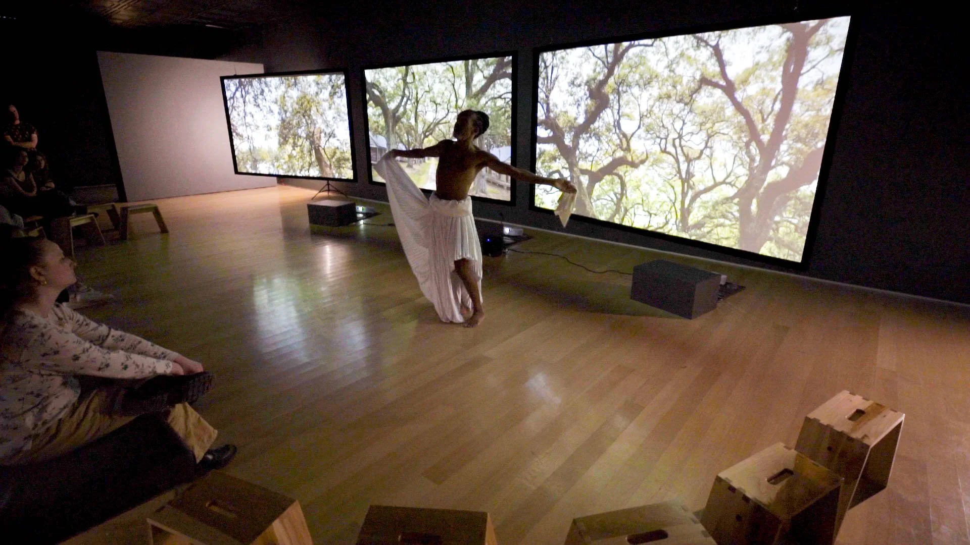 A woman performing a dance in front of three large screens displaying tree branches and leaves, with a small audience watching in a dimly lit room.
