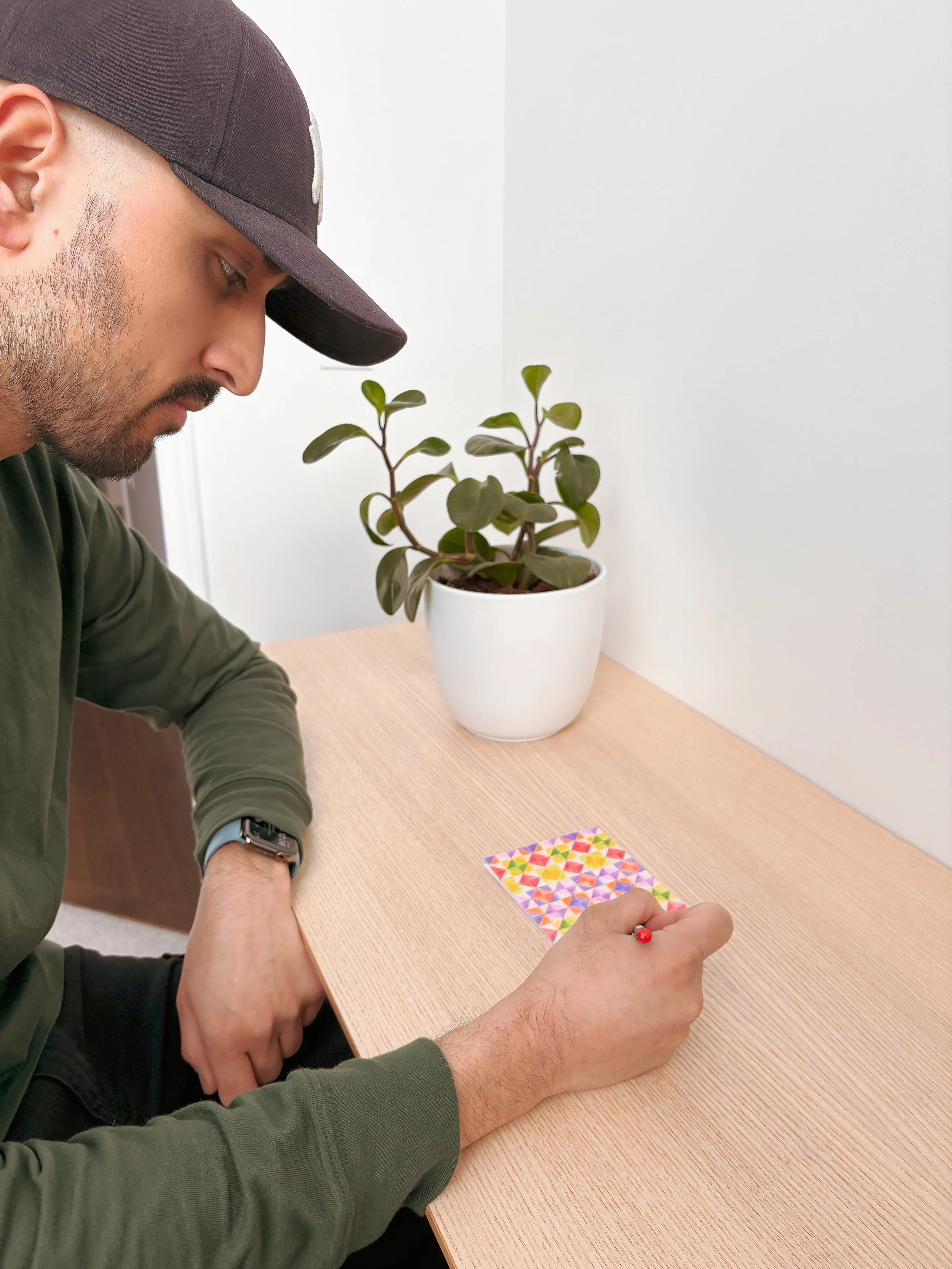 A man wearing a dark baseball cap and an olive green long-sleeve shirt is sitting at a light wood table, holding a red pen and drawing on a colorful geometric patterned paper. There is a potted plant with broad, dark green leaves in a white pot on the table, positioned against a plain white wall.