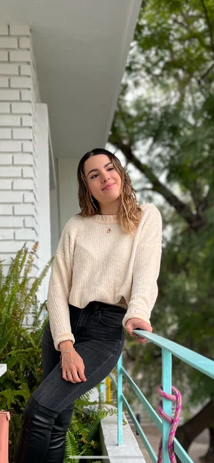Persona con suéter beige y pantalones negros posa en un balcón con plantas y barandilla azul.