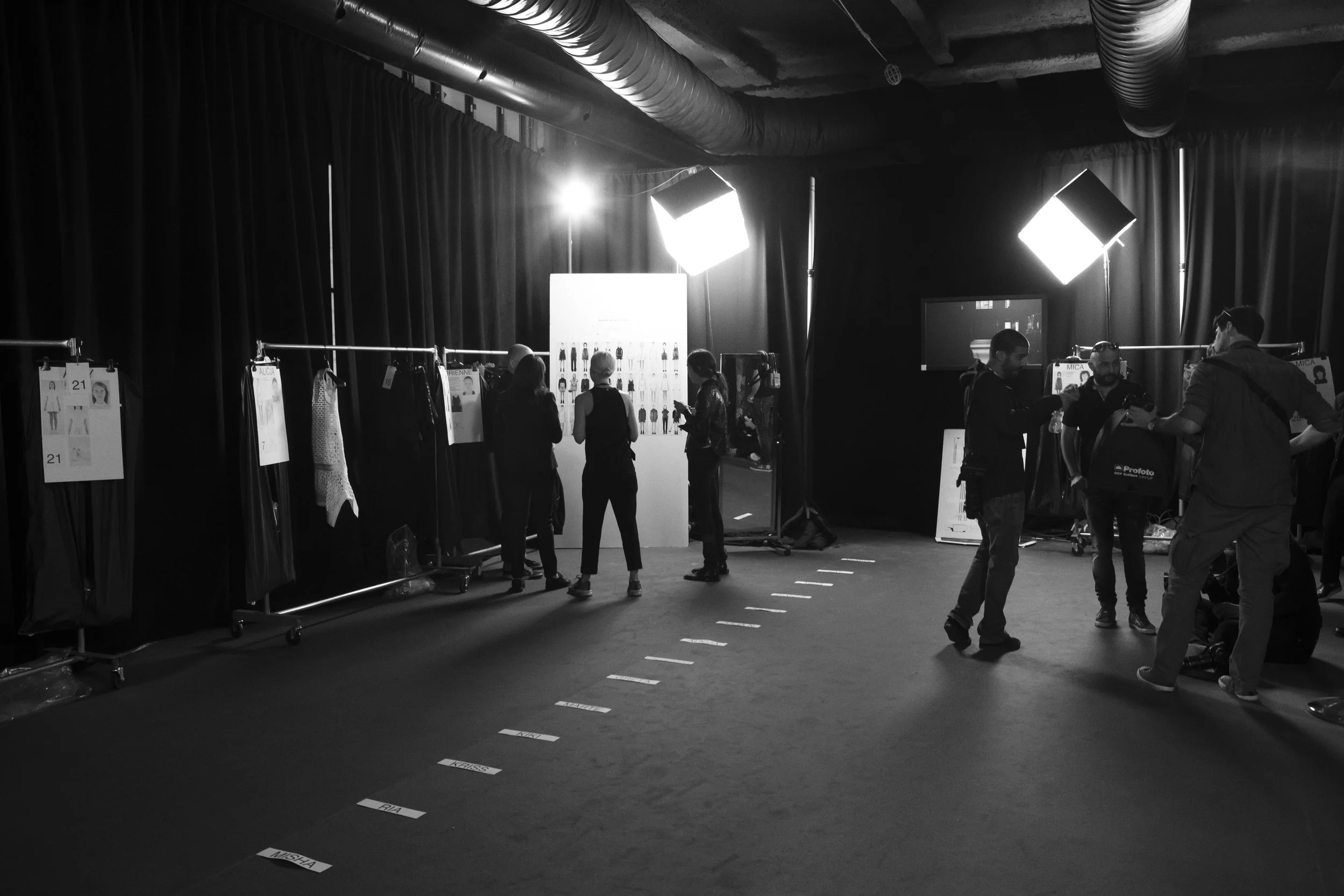 Personas trabajando en el backstage de un evento de moda, con estantes de ropa y luces de estudio.