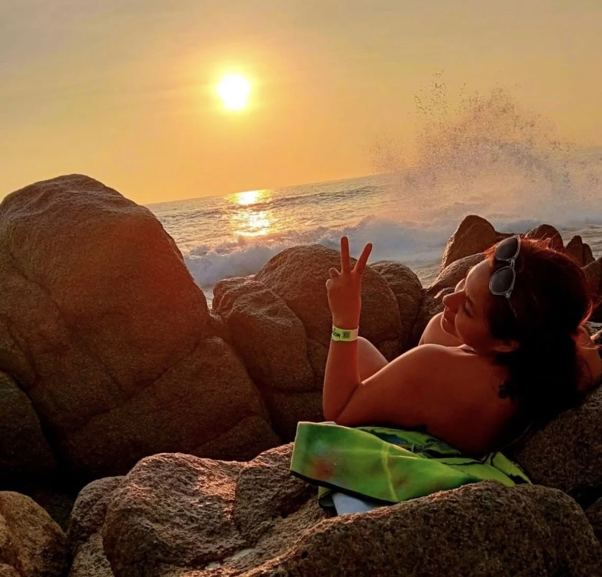 Persona relajándose en las rocas durante el atardecer en la playa, con el sol reflejándose en el mar y ola rompiendo en el fondo.