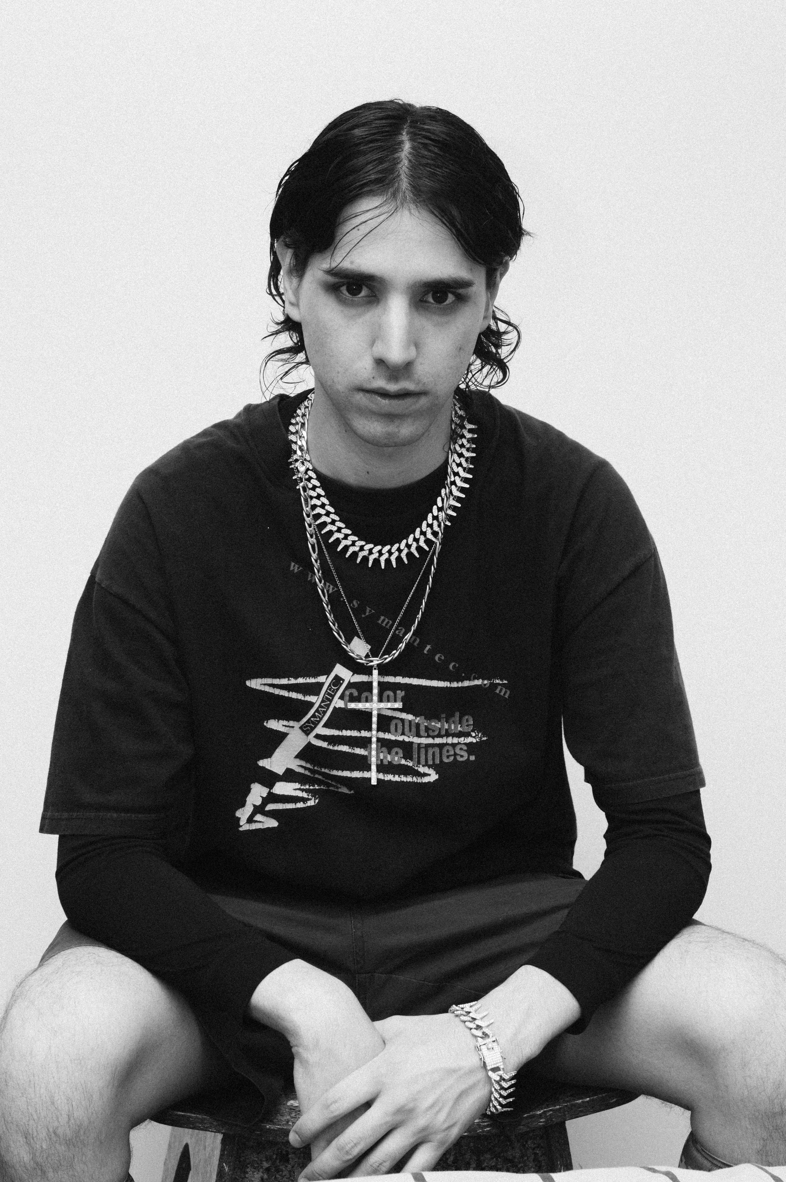 Retrato en blanco y negro de una persona joven con cabello oscuro, vistiendo una camiseta con diseño gráfico y collares.