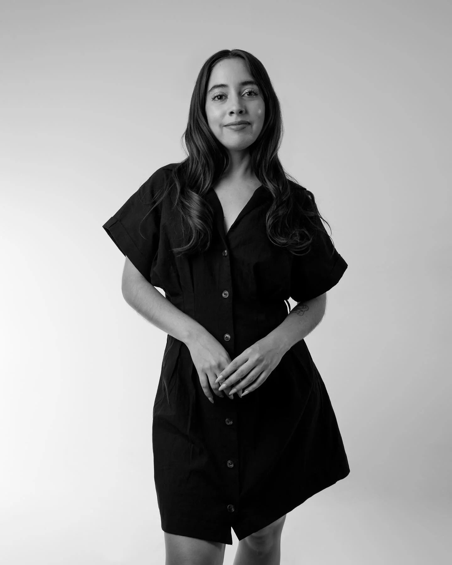 Mujer joven de pie en vestido negro, con fondo blanco, en una imagen en blanco y negro.