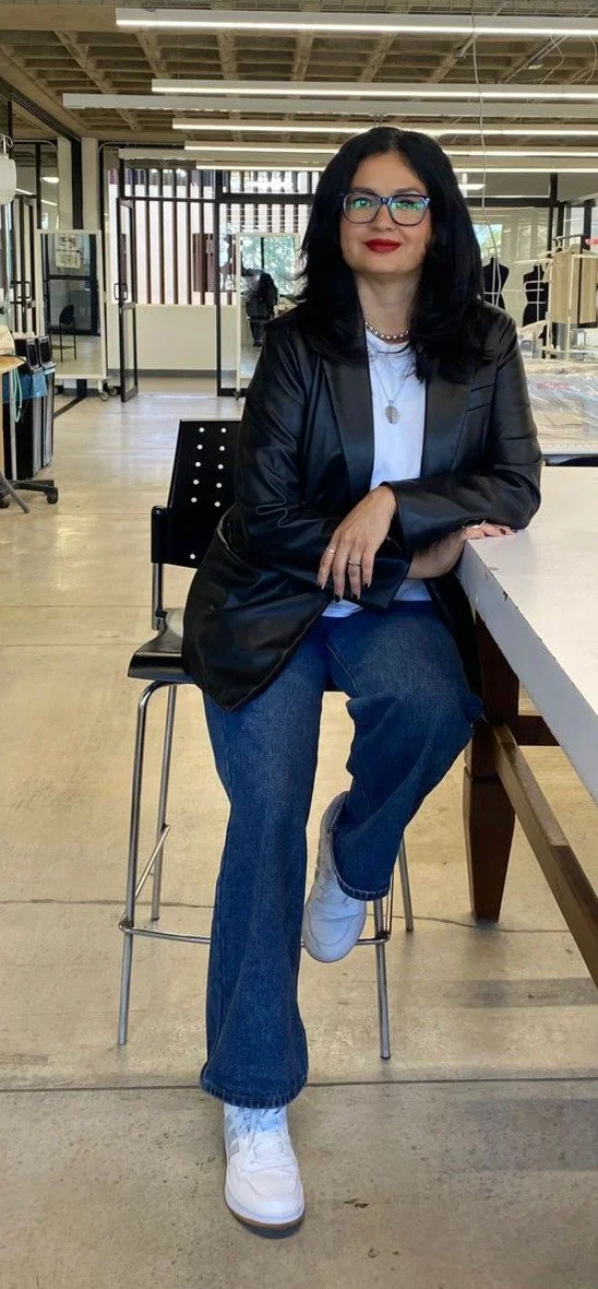 Persona sentada en un taburete dentro de una oficina moderna y luminosa. Lleva chaqueta de cuero negra, camiseta blanca, jeans azules y zapatos deportivos blancos. De fondo se ven mesas y equipos de trabajo.