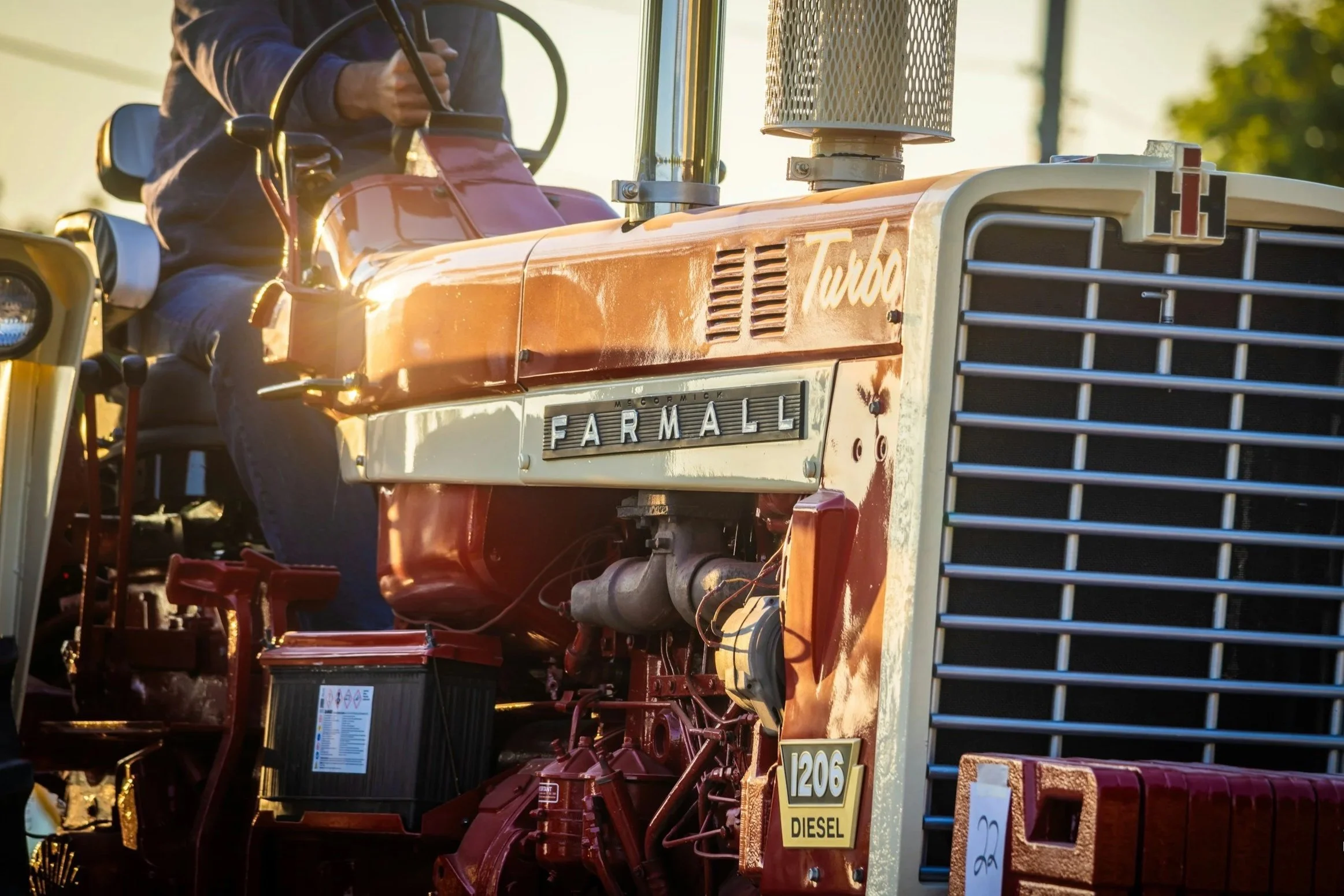 Close-up of a vintage Farmall tractor with a person sitting on it, sunlight highlighting its red and cream paint, showcasing details like the large front grille, a
