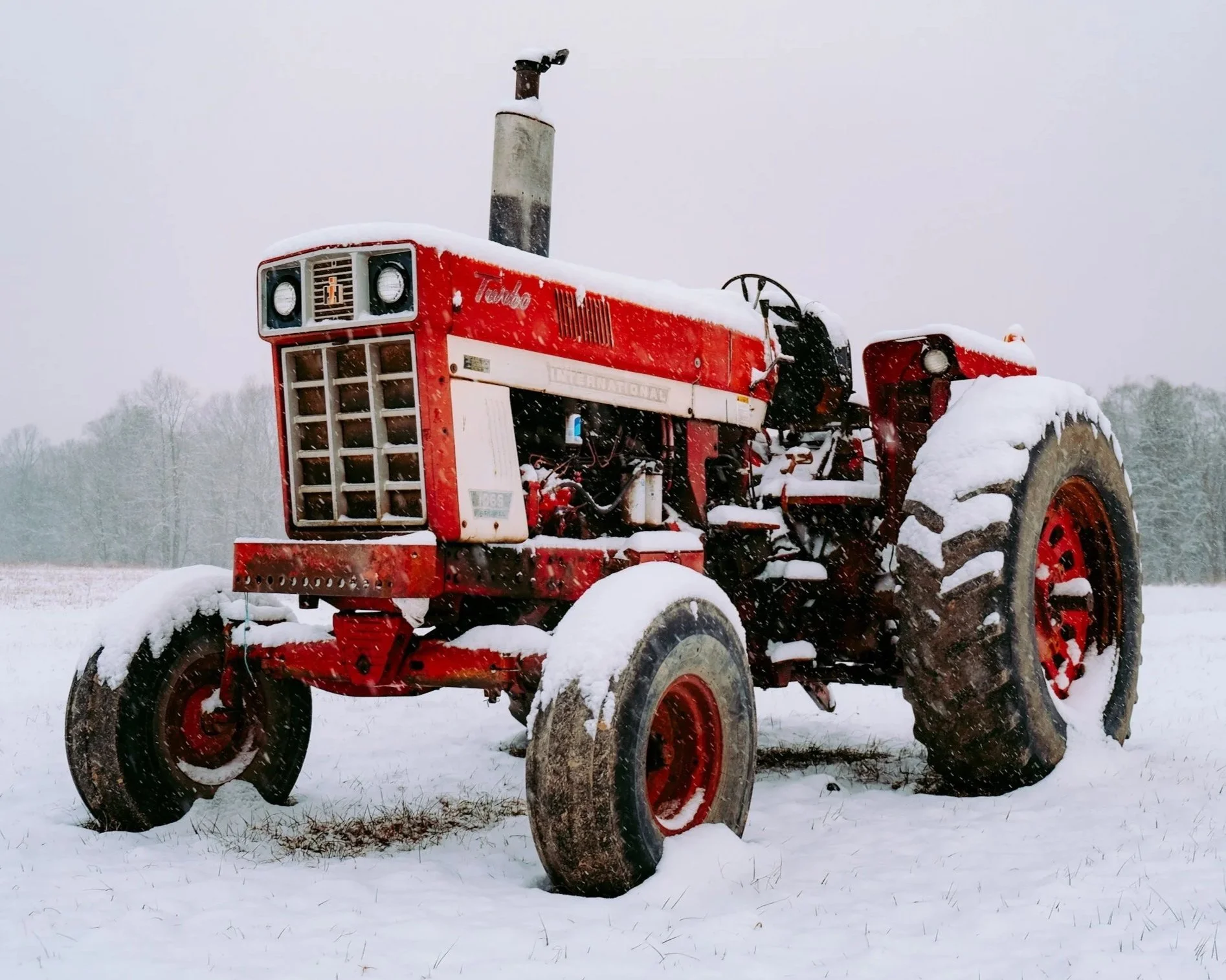 Snow-covered red tractor in a field with snow on its tires and body, and a background of snow and trees.
