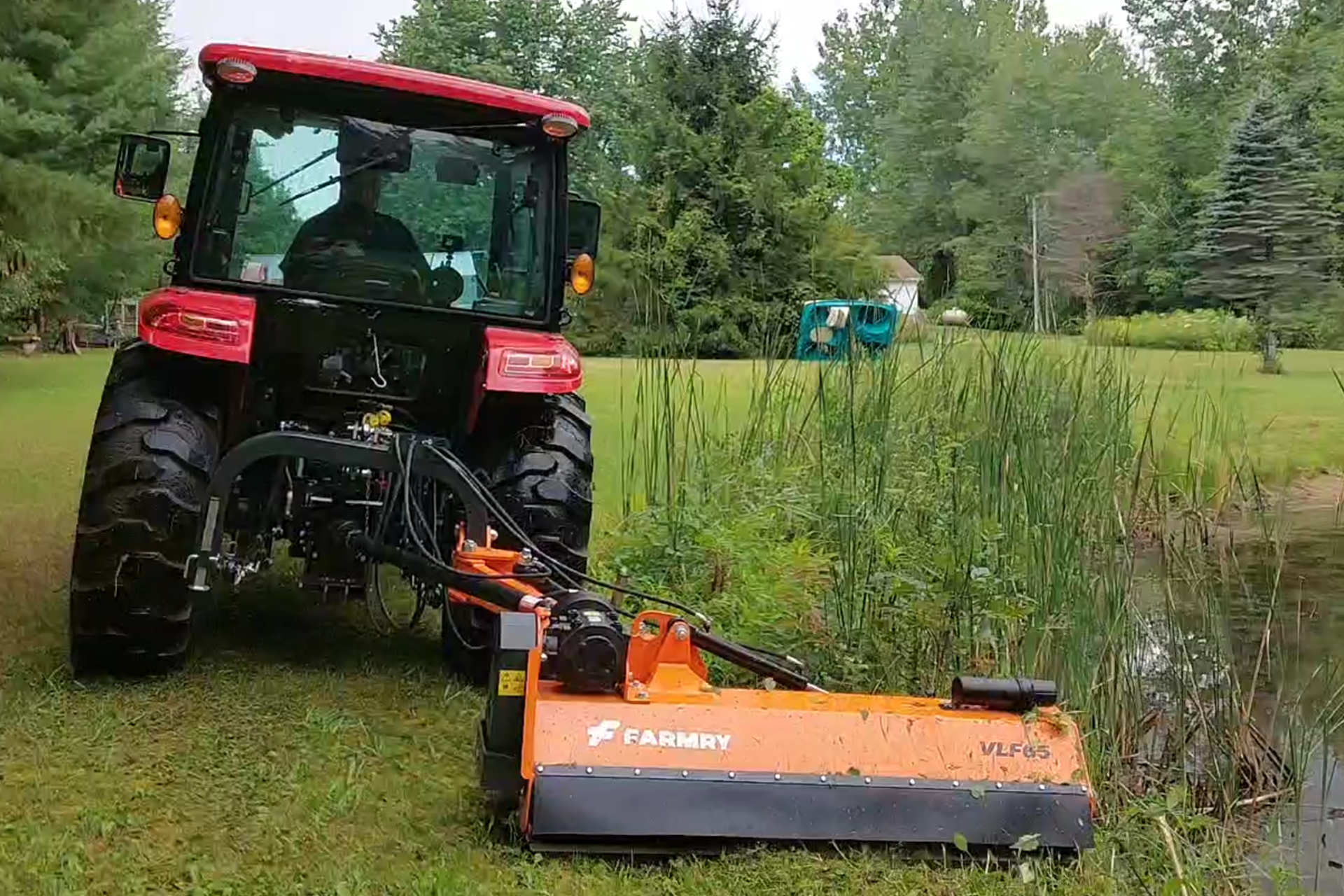 A red tractor with a mower attachment on a grassy area near a pond, with trees and a house in the background.