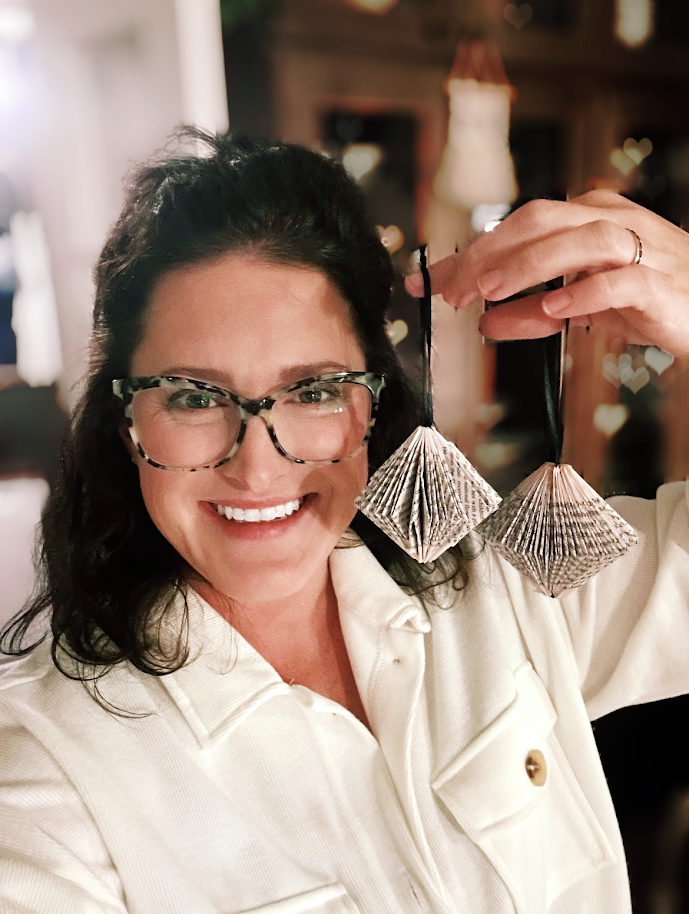 Woman with glasses smiling and holding two silver paper ornaments in a cozy, warmly lit room.