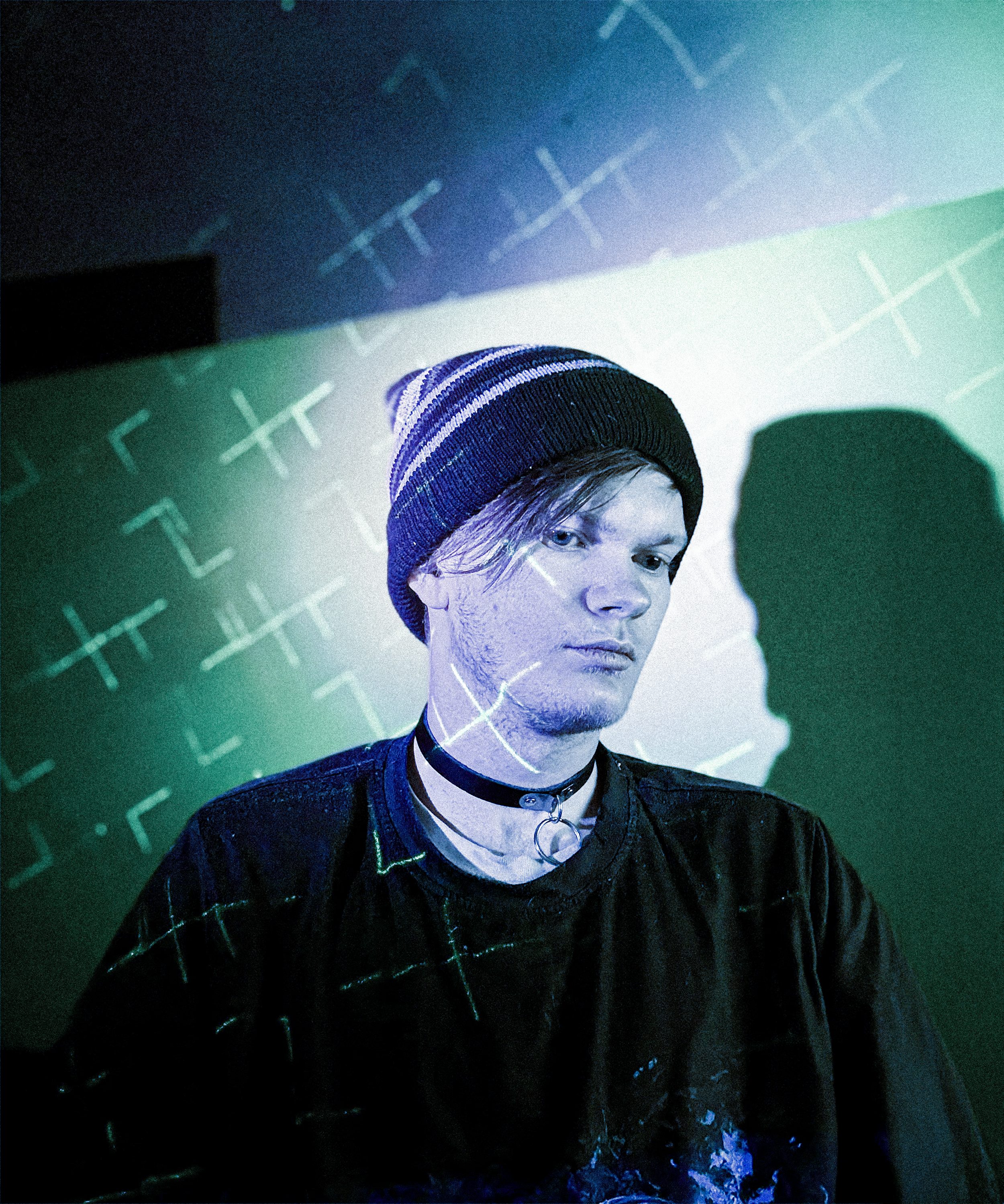 A young person with pale skin, wearing a striped beanie hat, a choker necklace, and a black shirt with paint splatters, looks to the side with a serious expression. The background features a projection of abstract lines and symbols.