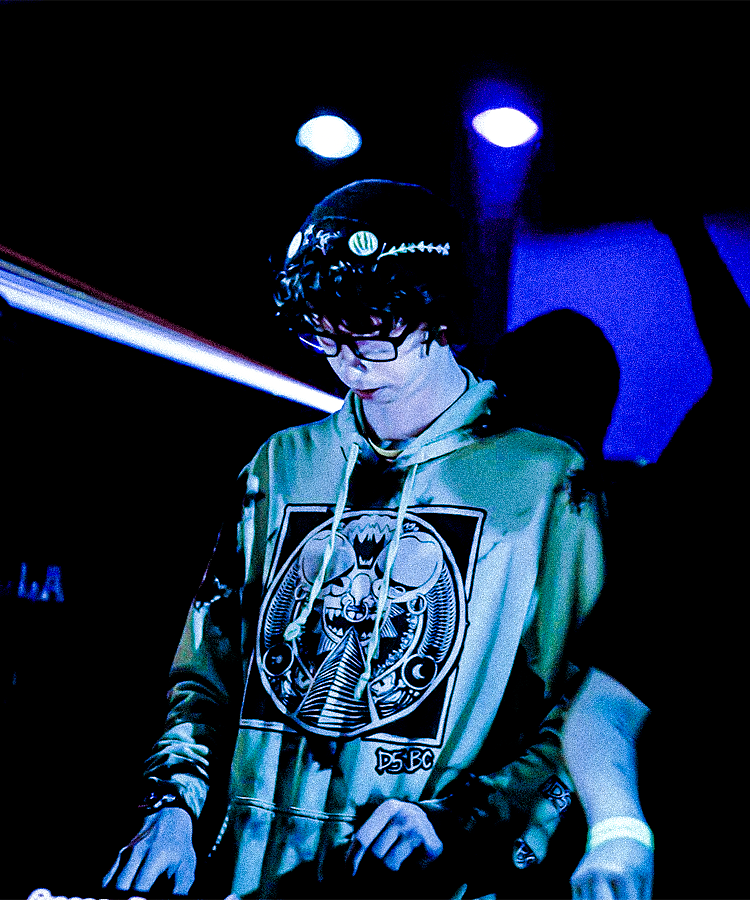 Person with curly hair, glasses, and a beanie hat, wearing a graphic hoodie, in a dark room with blue lighting.